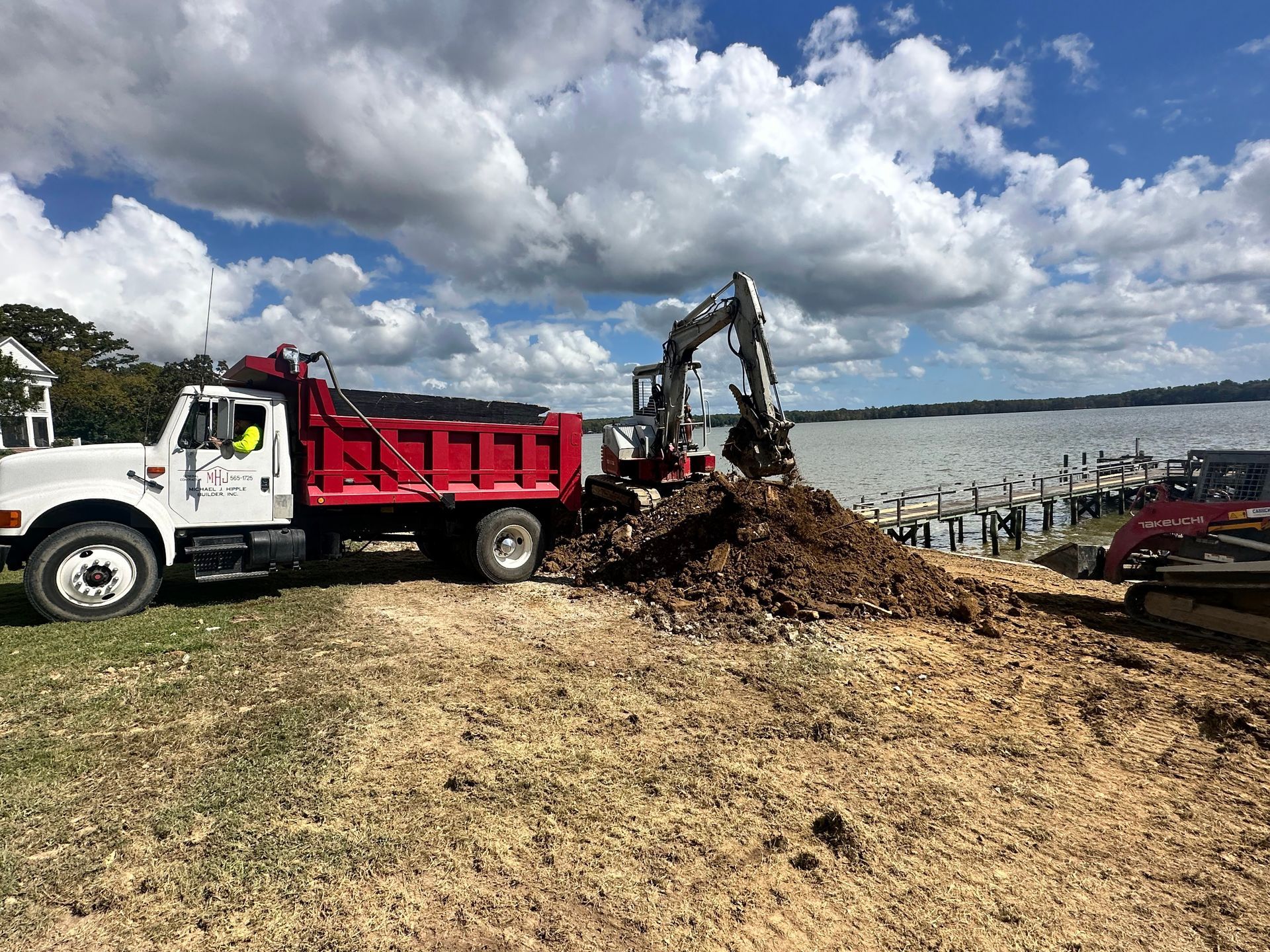 A dump truck and excavator work on a waterfront site under a cloudy sky.