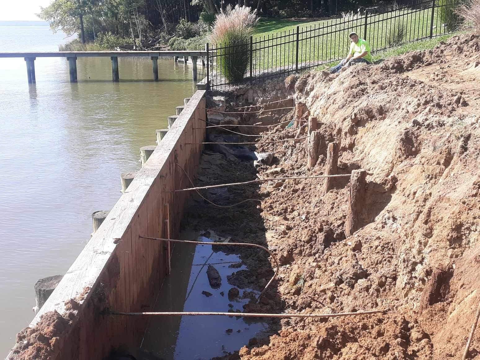 Construction of a retaining wall on a lake. Brown soil, wooden posts, metal rods, and water visible.