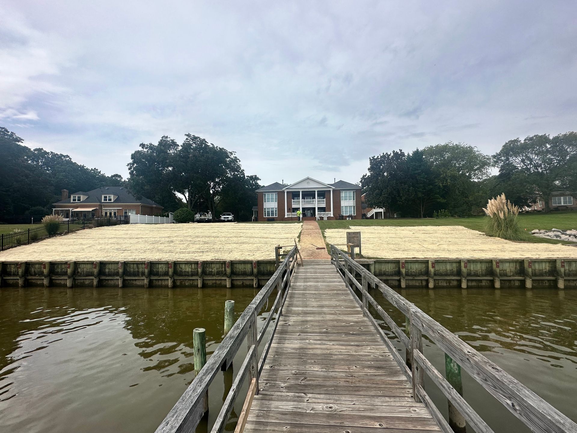 Wooden pier leads to a large white house with columns, flanked by grassy areas, near a body of water.