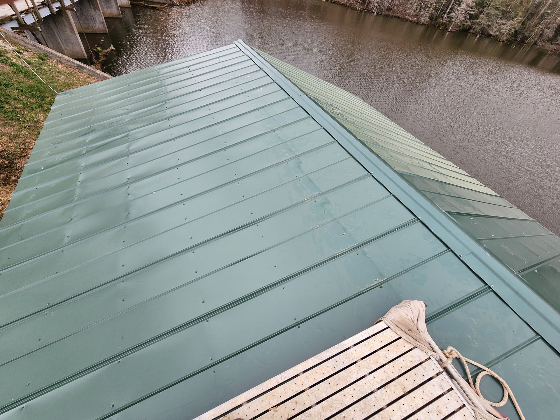 A high-angle view of a green metal roof sloping toward a river, with a portion of a white bench in the foreground.