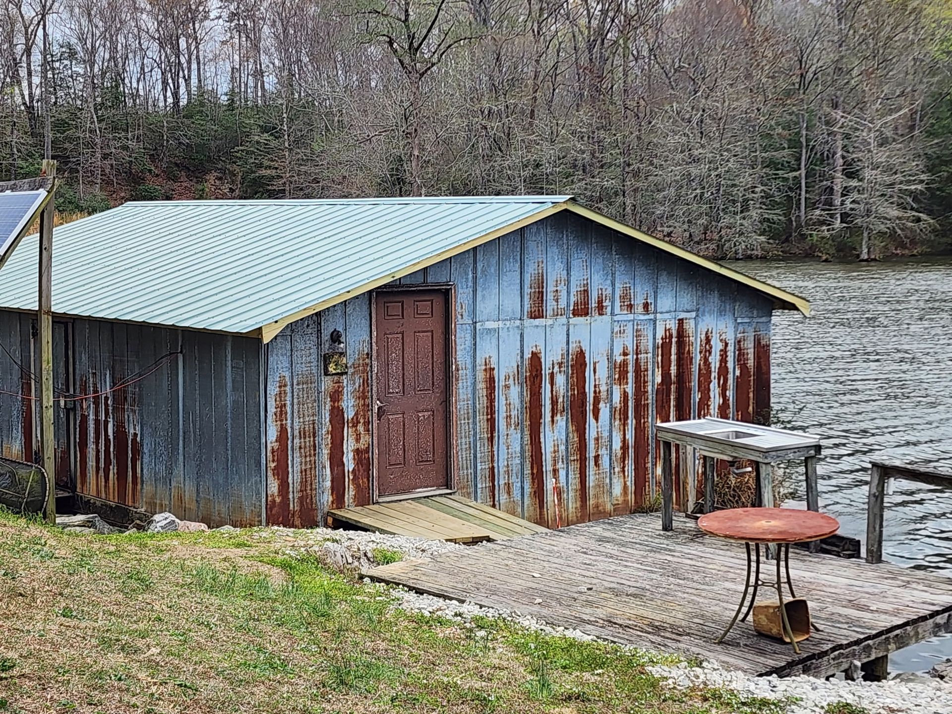 A weathered, rusted metal boat house sits on the edge of a lake, featuring a brown door and a small outdoor patio.