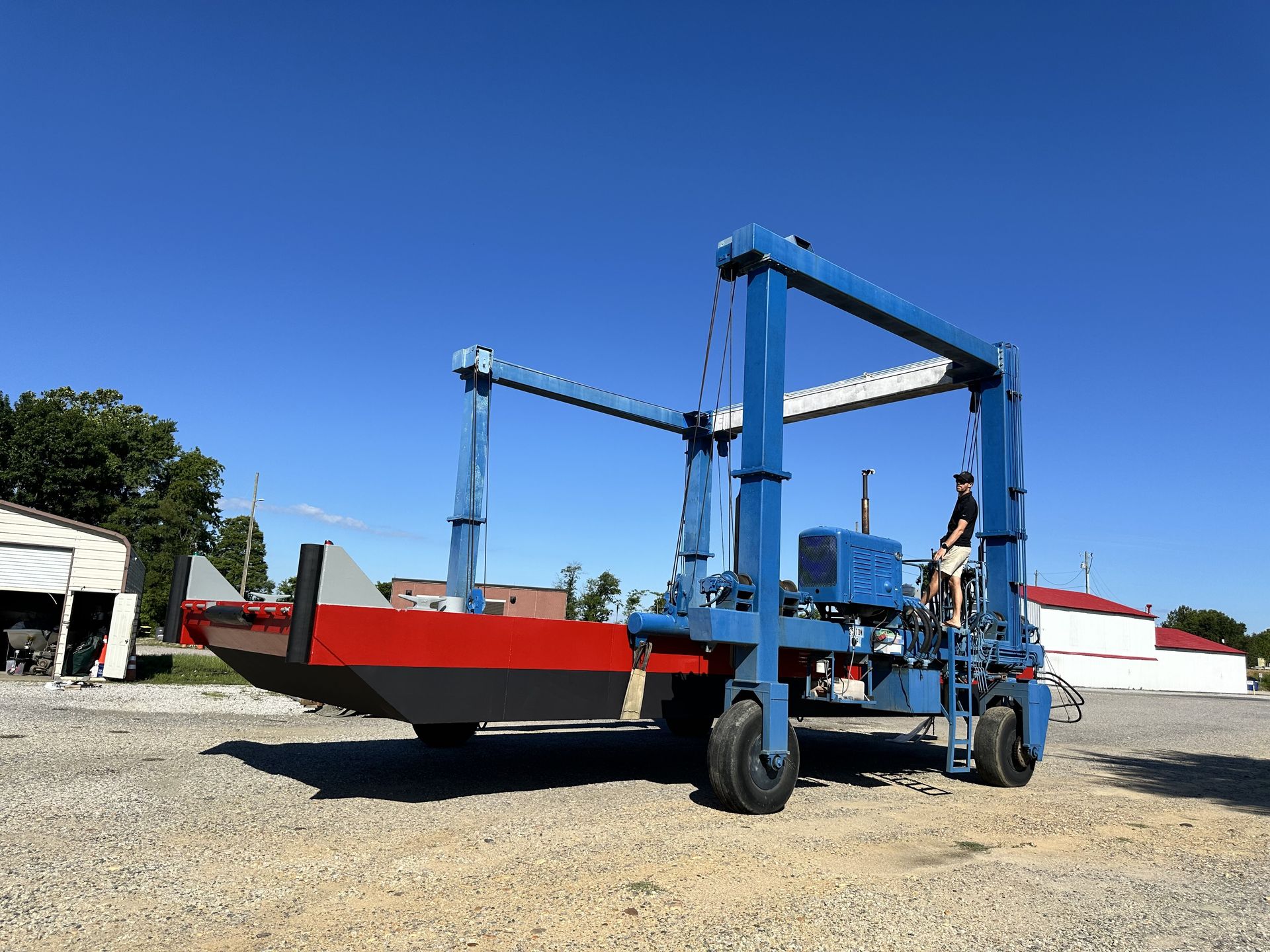 Blue boat lift holding a red and gray boat on gravel under a blue sky.
