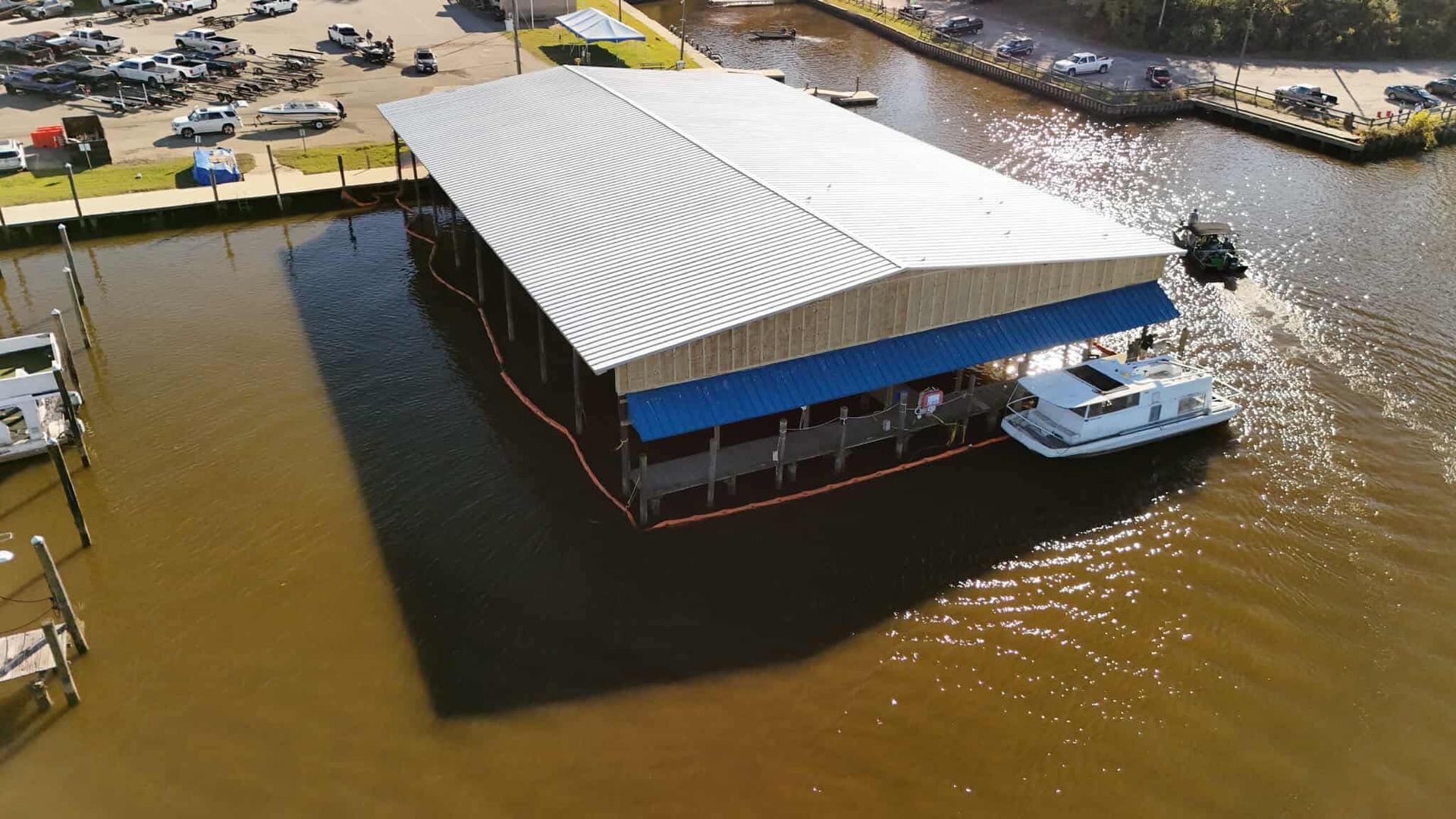 Boat under a large covered dock on the water, surrounded by docks, vehicles, and the shore.