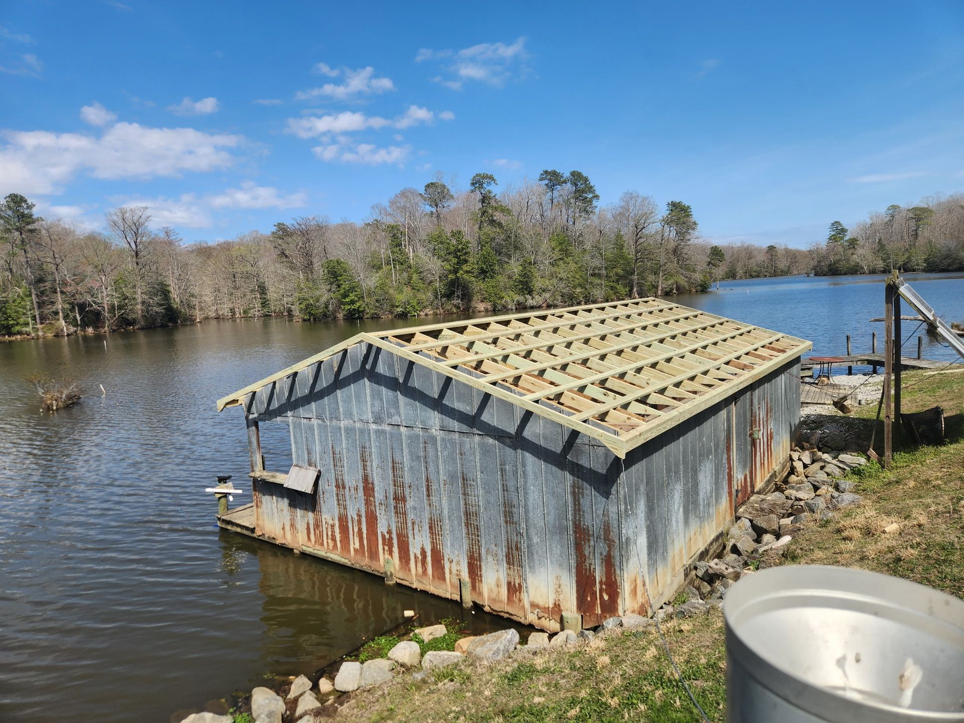 A rusted, shed-like boat house with an exposed wooden frame roof sits partially submerged in a calm lake on a sunny day.