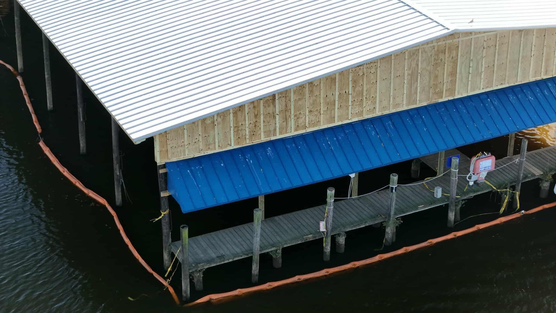 A wooden boathouse with a blue roof, built on a pier, surrounded by water. An orange barrier floats nearby.