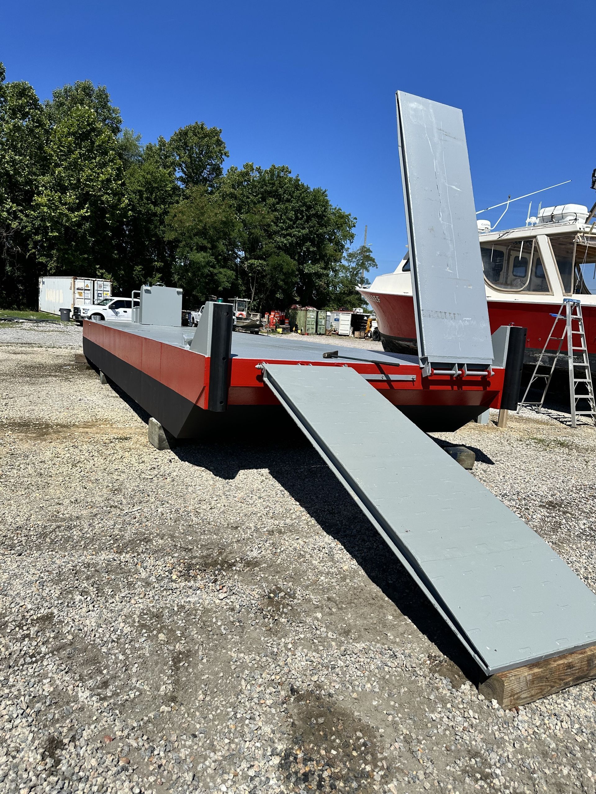 A red and black barge with a gray ramp on a gravel surface under a blue sky.