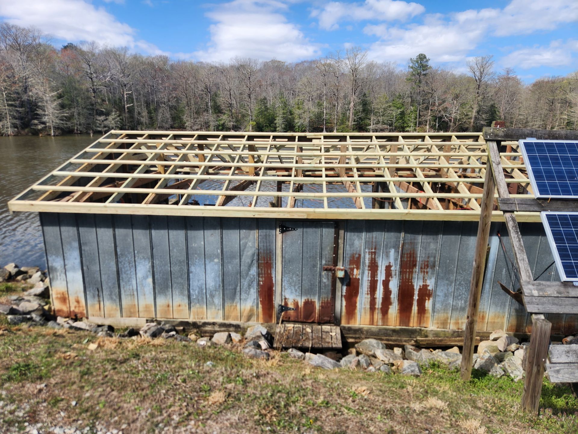 A rustic shed with rusted metal siding and a newly constructed wooden roof frame, located near a lake with solar panels.