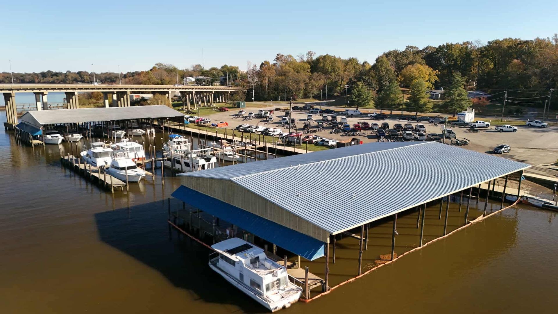 Boats docked at a marina under metal-roofed covers; cars parked nearby; bridge in the background.