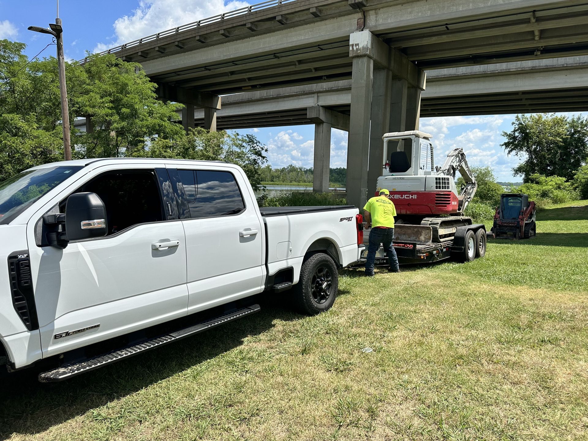White truck with a trailer carrying an excavator under an overpass, person is working on the trailer.