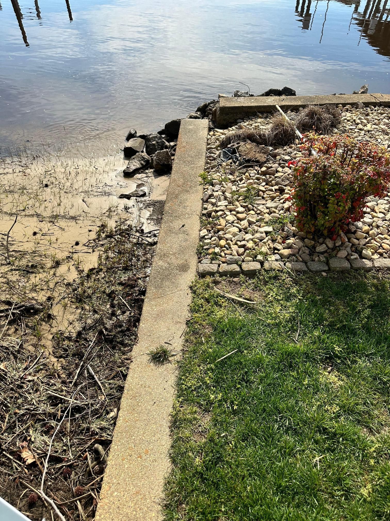 A concrete edge separates water and landscaping, featuring pebbles, plants, and grass.