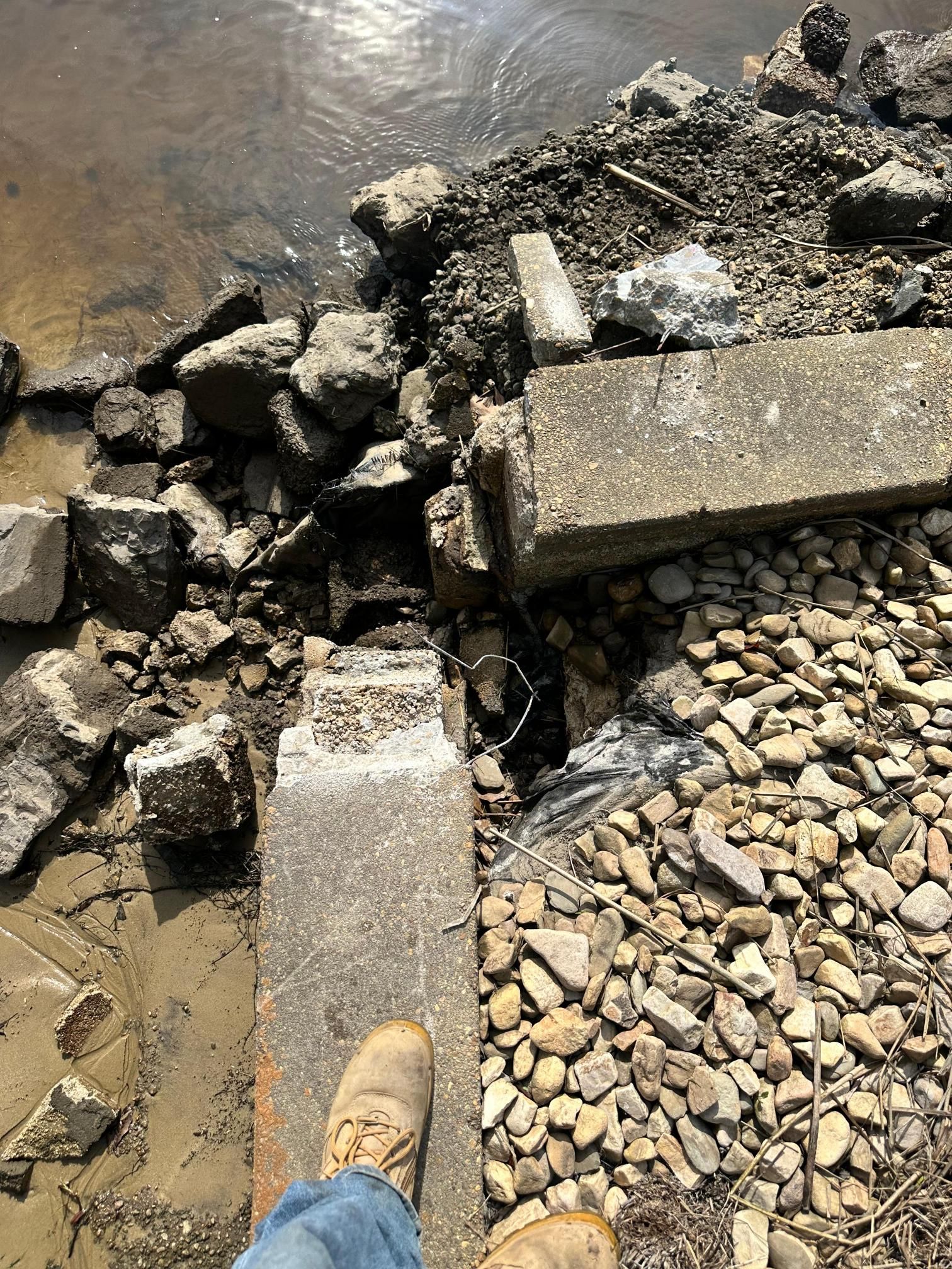 Person's foot near a dilapidated concrete edge with rubble, small rocks, and water nearby.