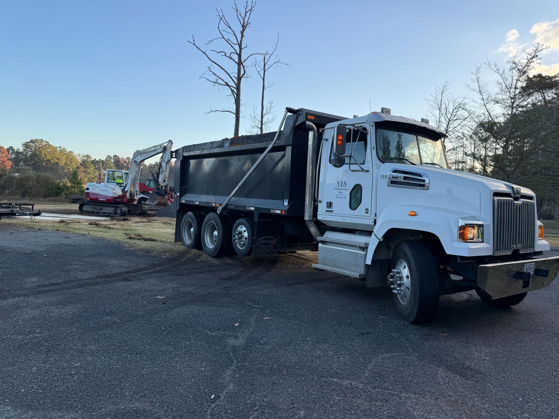 White dump truck and excavator on gravel in a wooded area under a blue sky.
