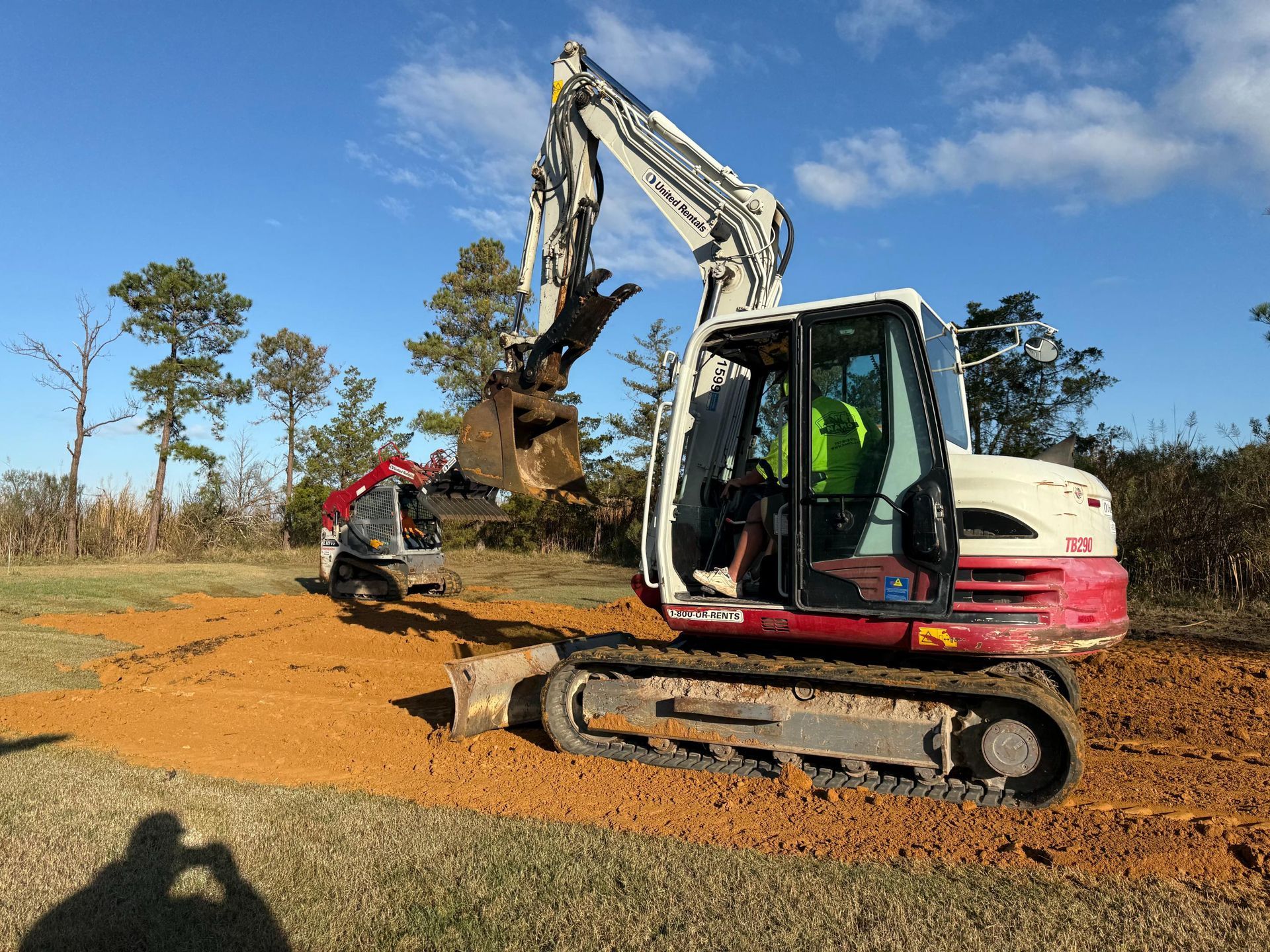 Excavator digging in dirt on a sunny day. A small red excavator is in the background.