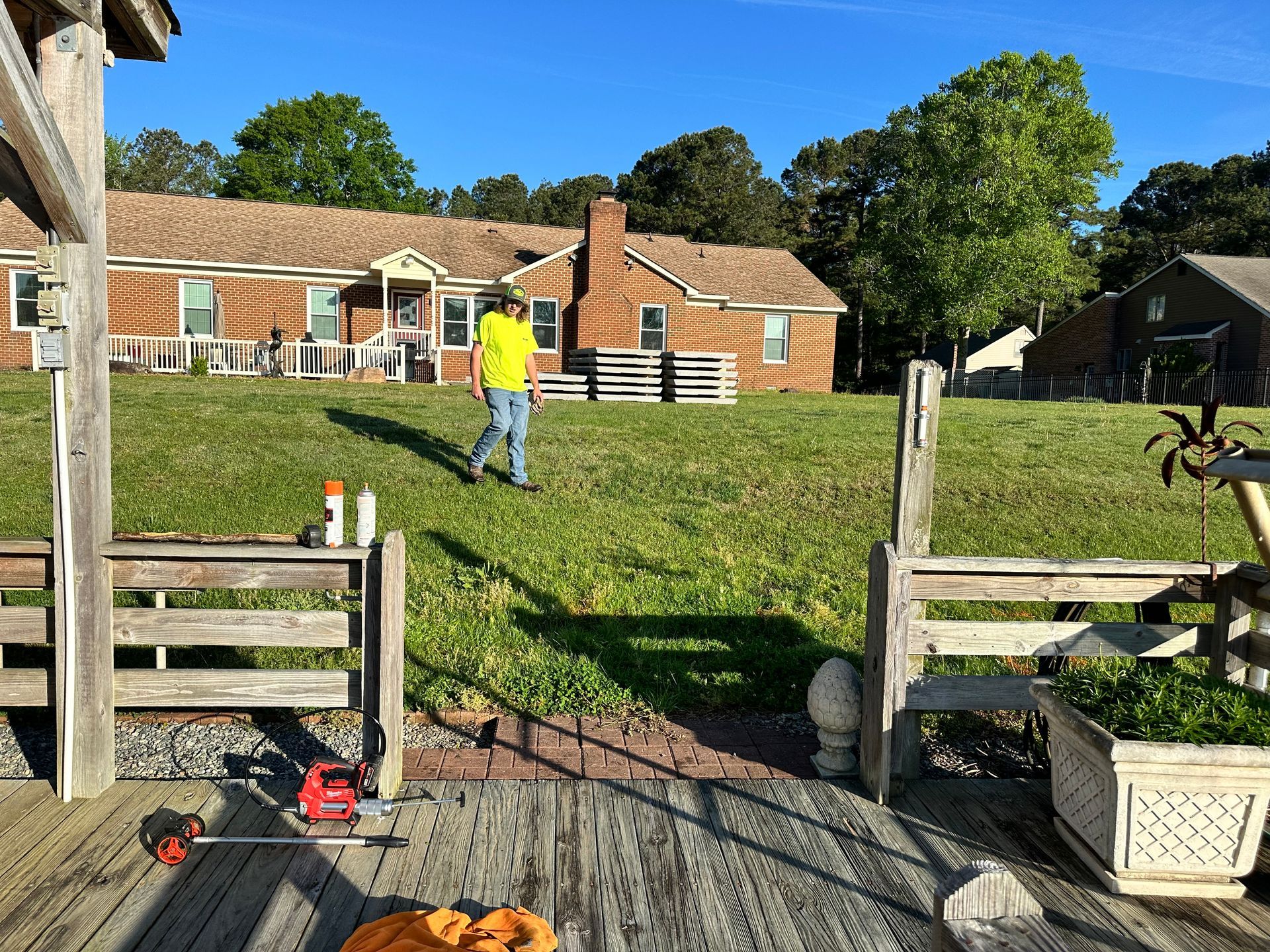 Man in yellow shirt walks on grassy yard towards a house. Wooden dock and tools in foreground.