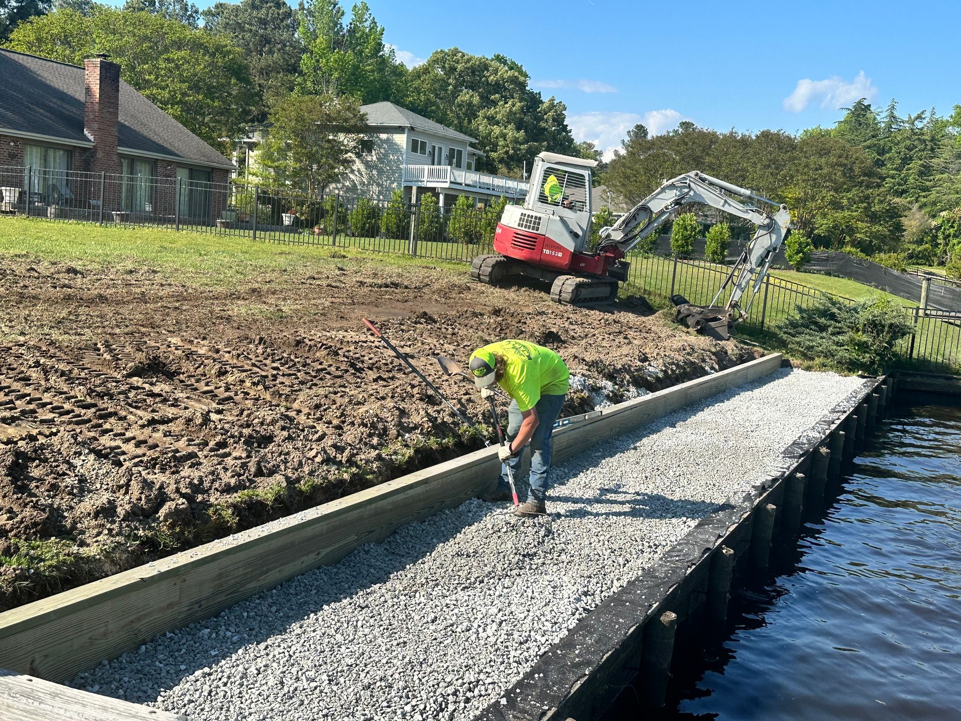 Construction workers installing gravel on a shoreline; excavator in background, houses on a hill.