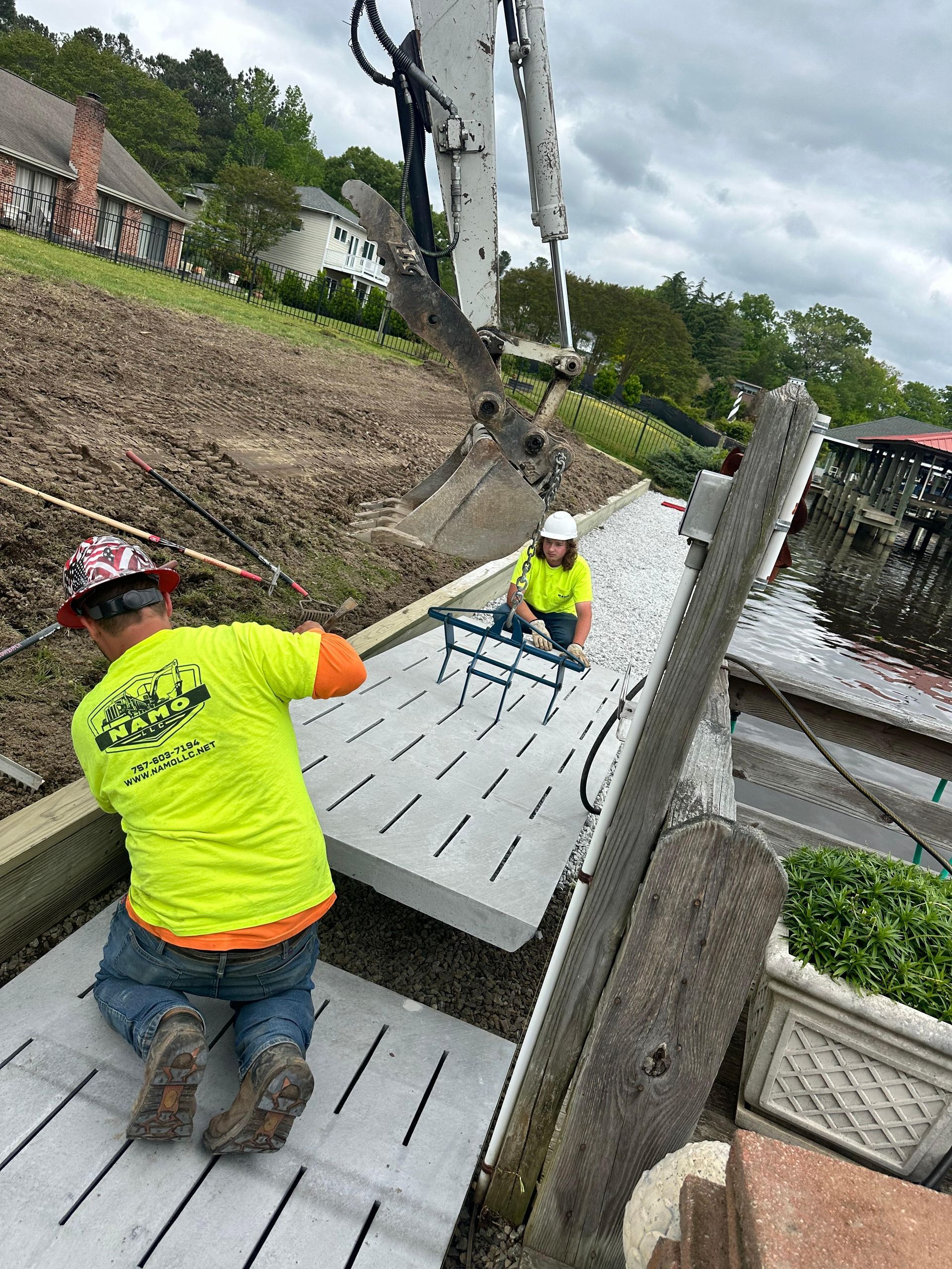 Two construction workers install a concrete walkway near water. One kneels, holding a slab. The other uses a tool.