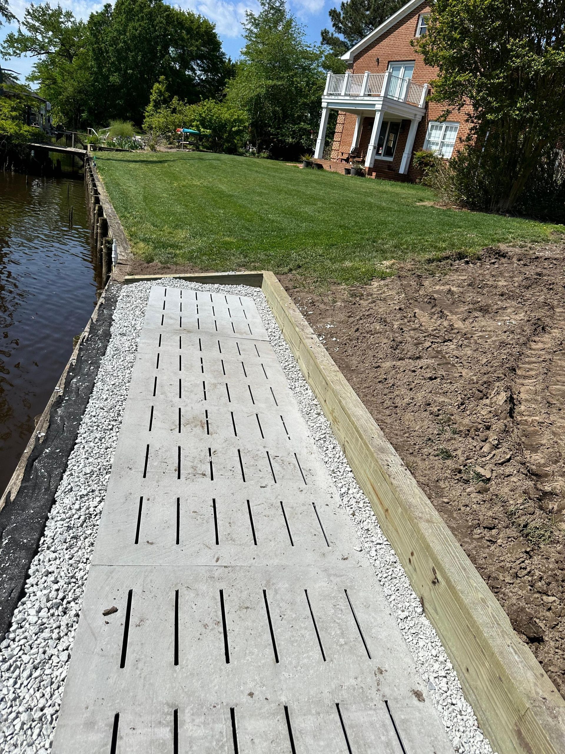 Concrete path with slotted design, bordering a body of water and lawn, next to a building and landscaping.