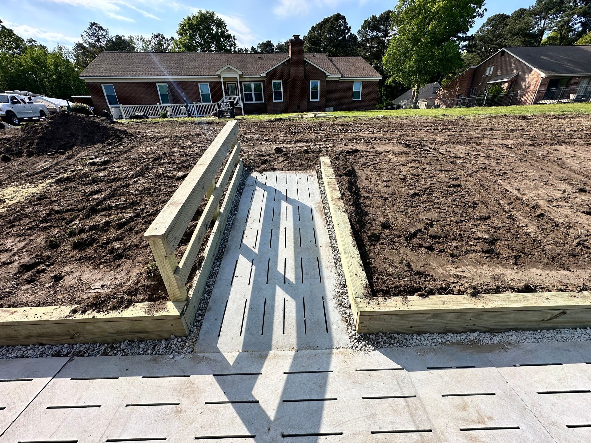 Wooden ramp with gray pavers leads to a house between soil beds.