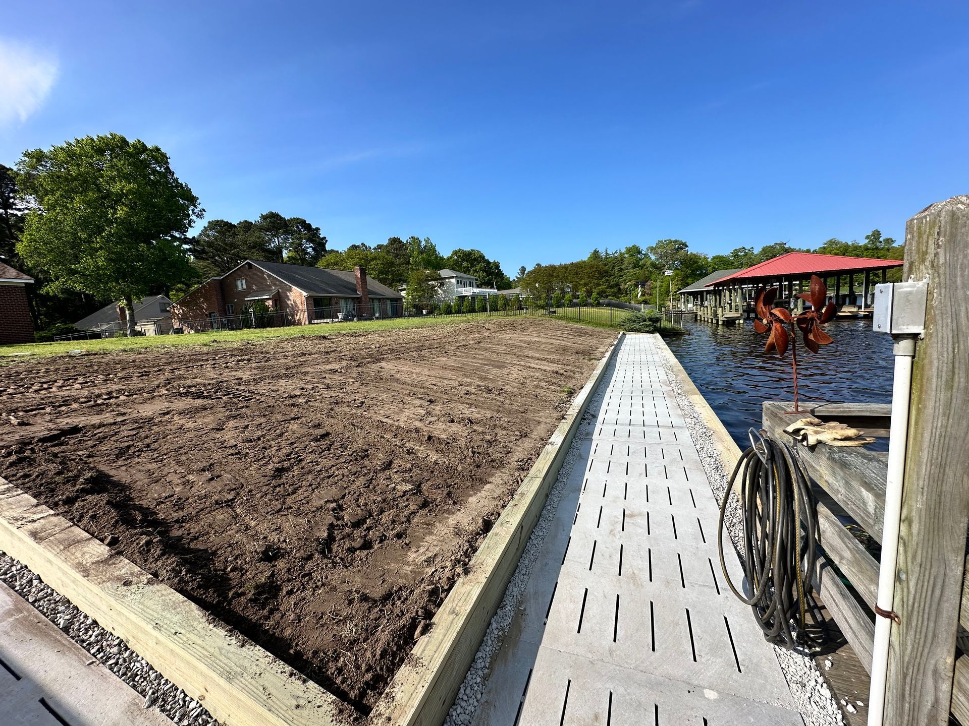 Dirt bed bordered by wood with walkway alongside water, houses and trees in background.