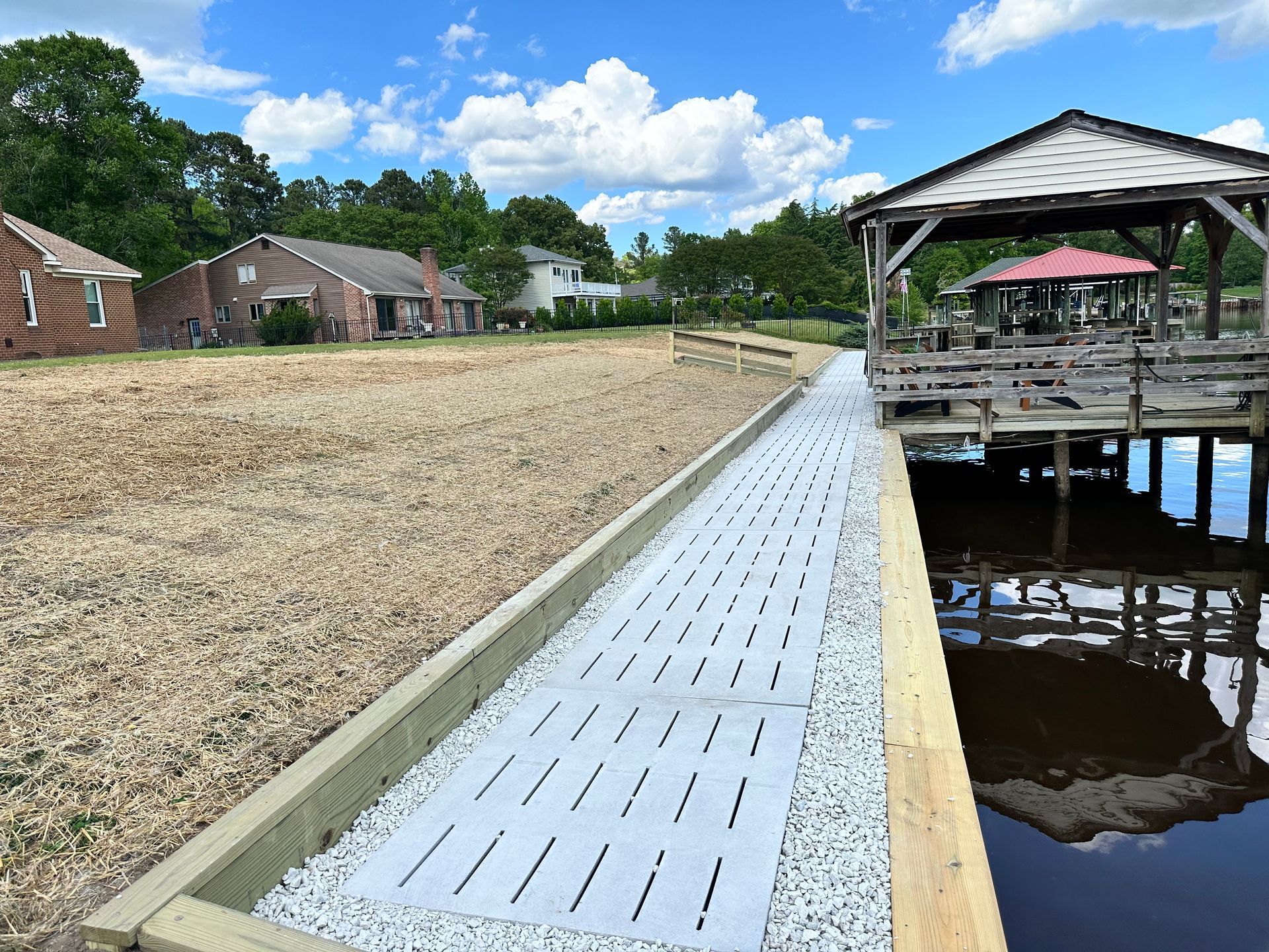 Path with grey pavers alongside water, leading to a wooden dock with a gazebo. Houses and trees in the background.