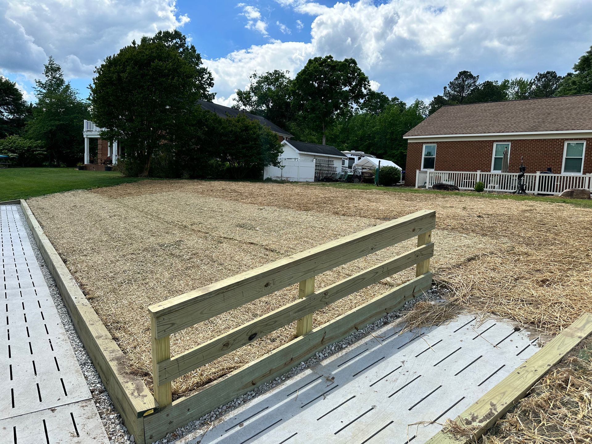 A rectangular area with wood chips, a wooden fence, and a concrete pathway; houses in the background.