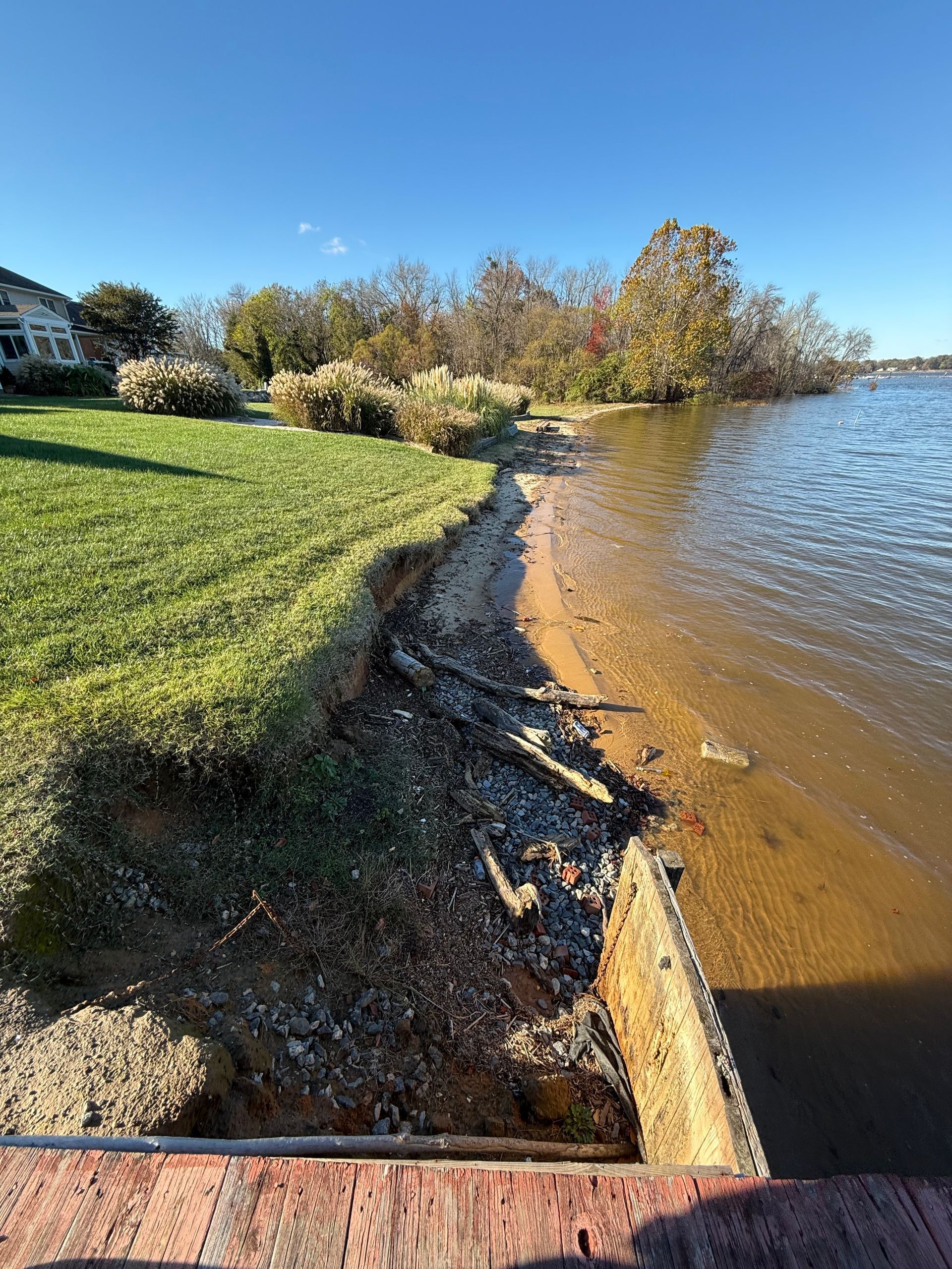 Lakeshore erosion; grassy lawn meets shoreline. Partially submerged concrete barrier, water, trees, and blue sky.