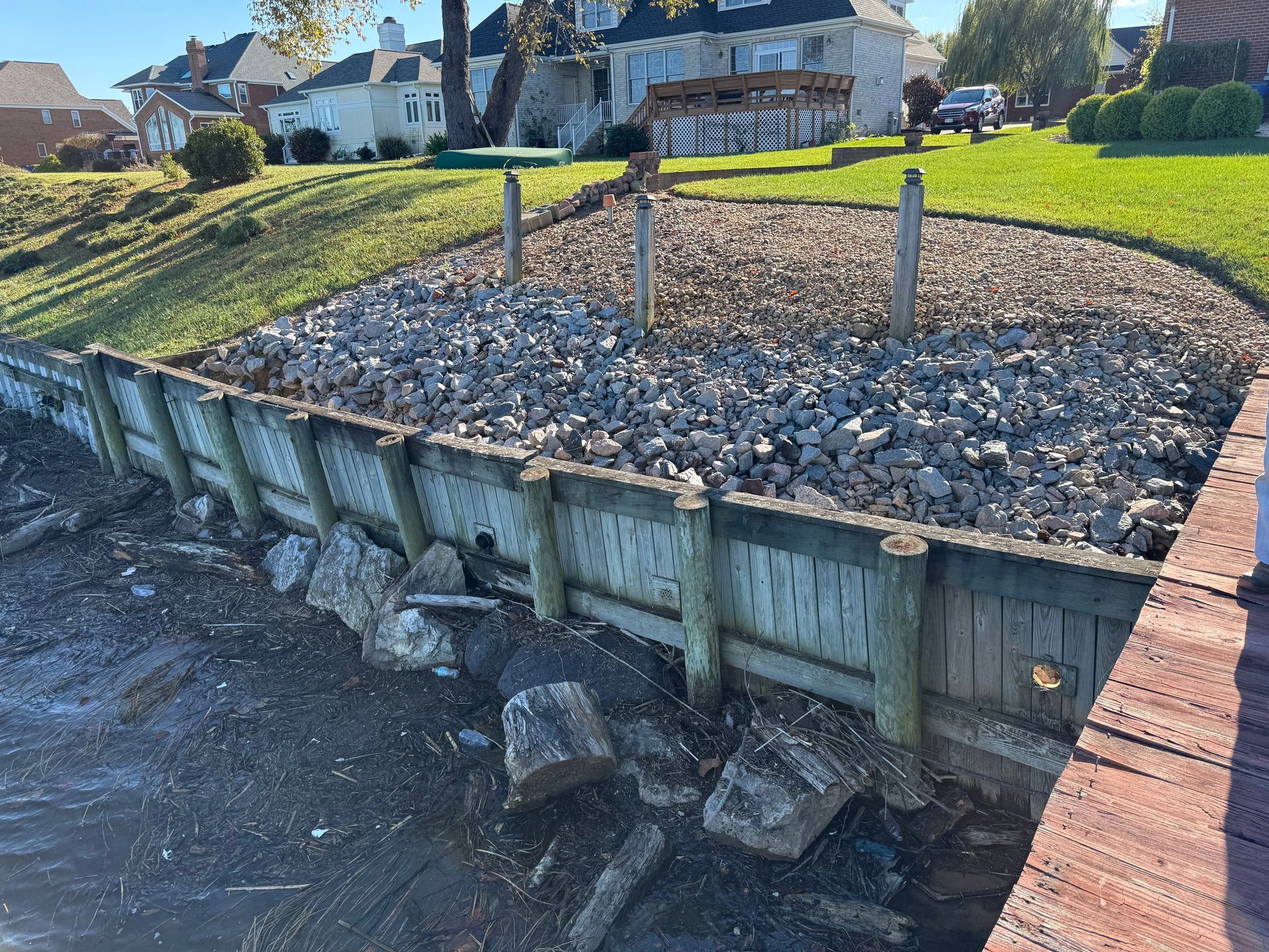Wooden retaining wall on a shore, filled with rocks, grassy hillside and houses in the background.