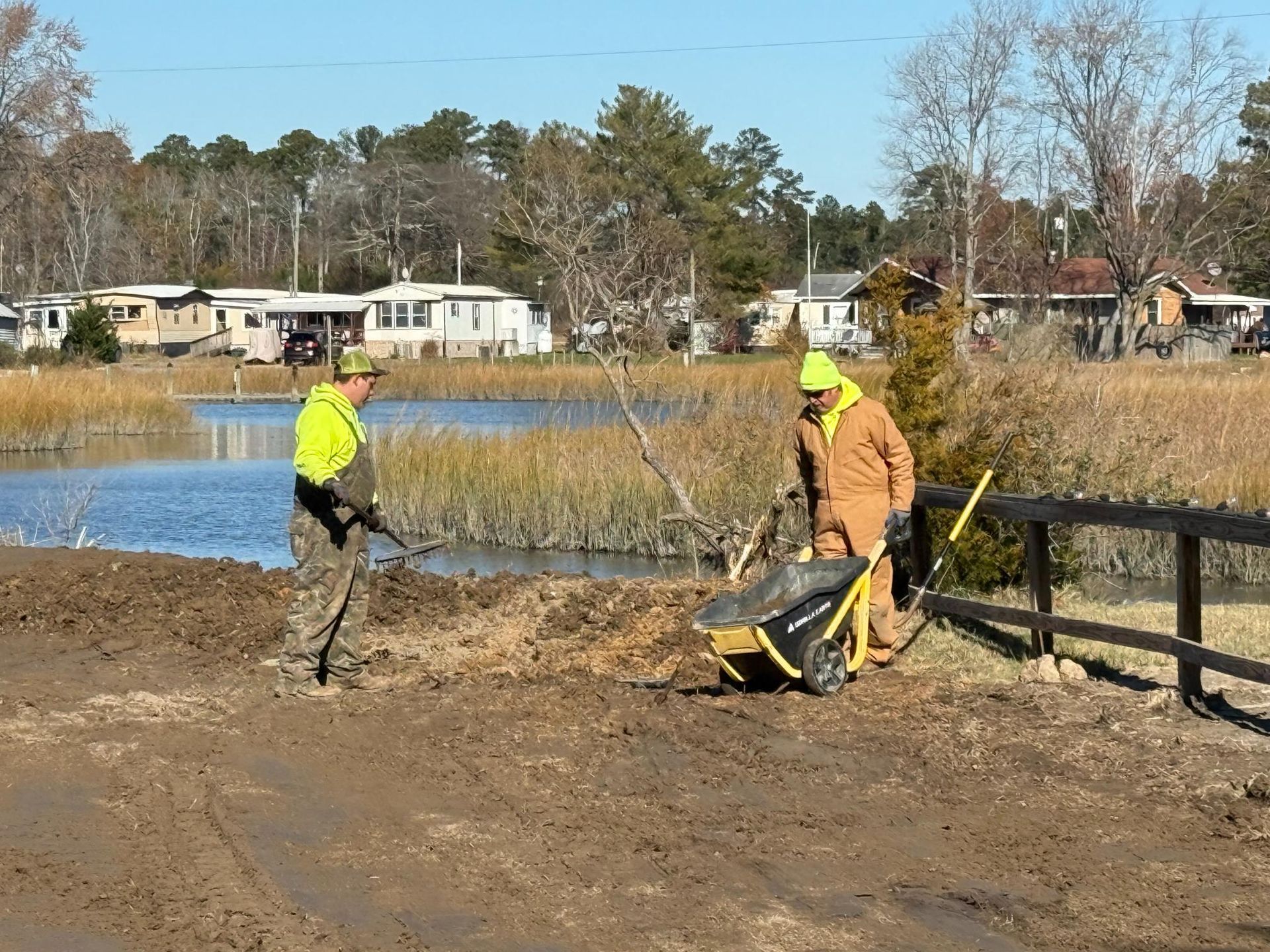 Two workers in high-vis vests and coveralls repair a muddy road near a lake, using a wheelbarrow and tools.