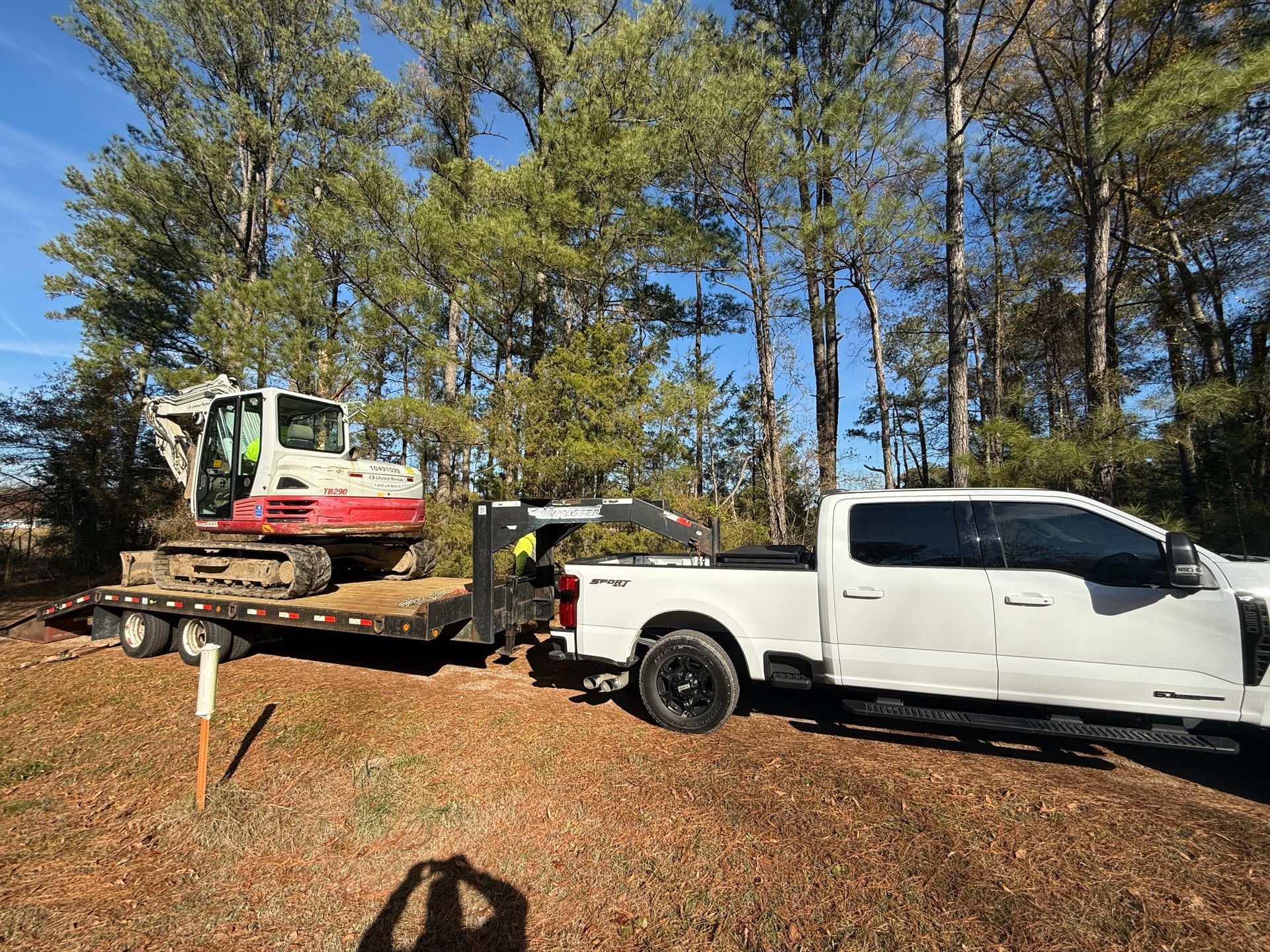 White truck towing an excavator on a trailer; outdoors, trees in background, sunny.