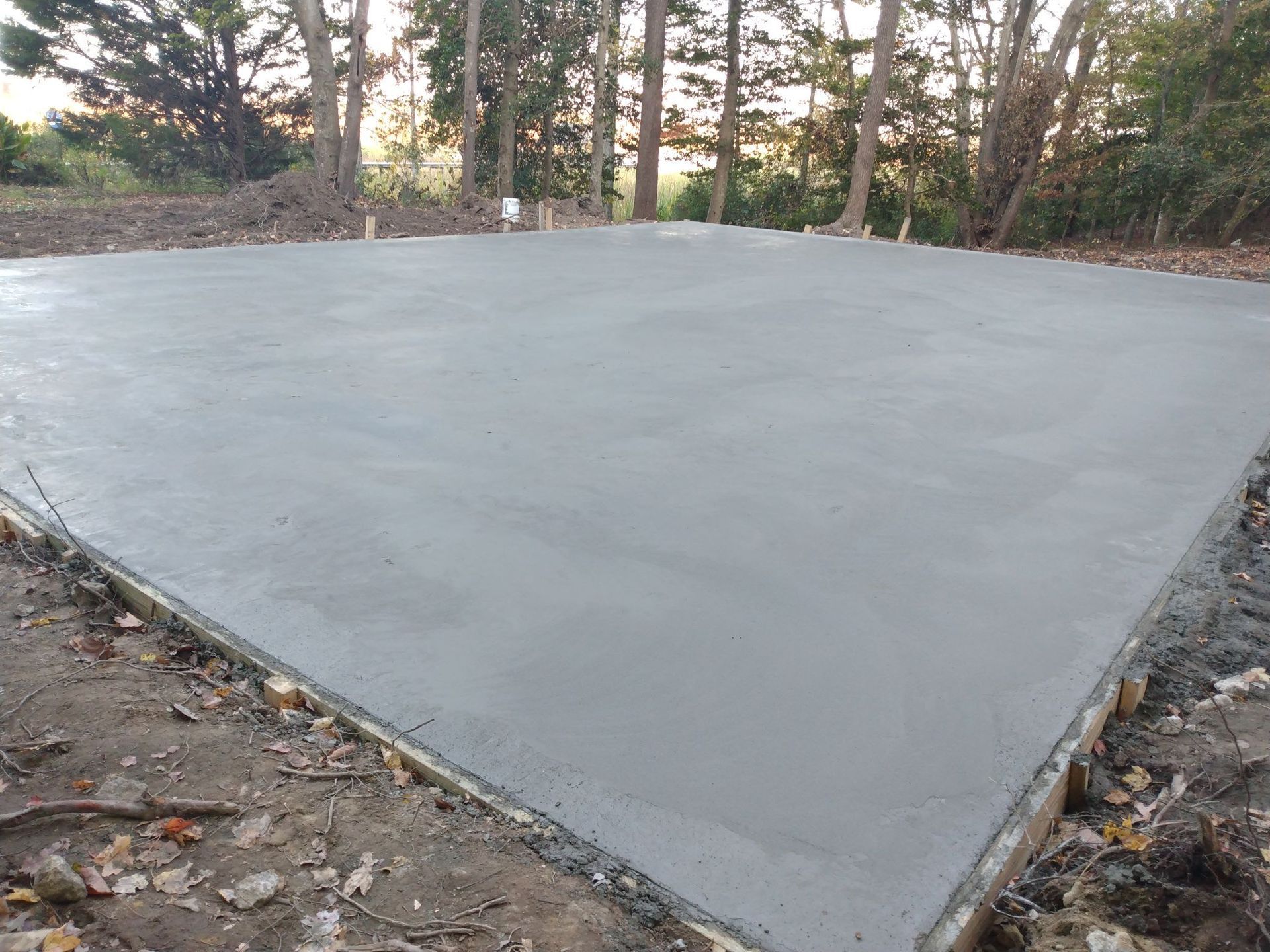 Newly poured concrete slab in an outdoor setting, surrounded by dirt and trees.