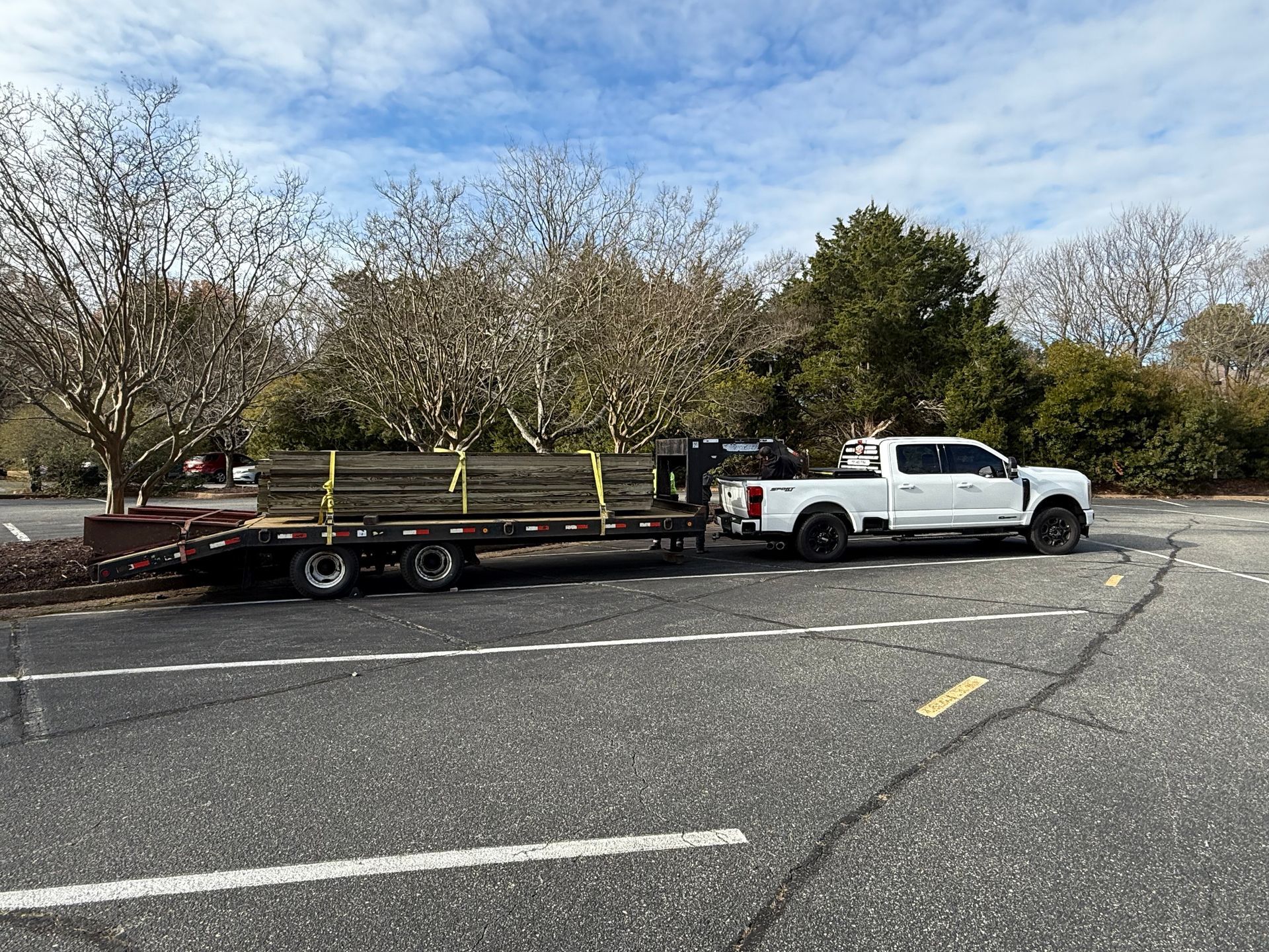 White truck towing a flatbed trailer loaded with lumber, parked in an asphalt parking lot under a cloudy sky.