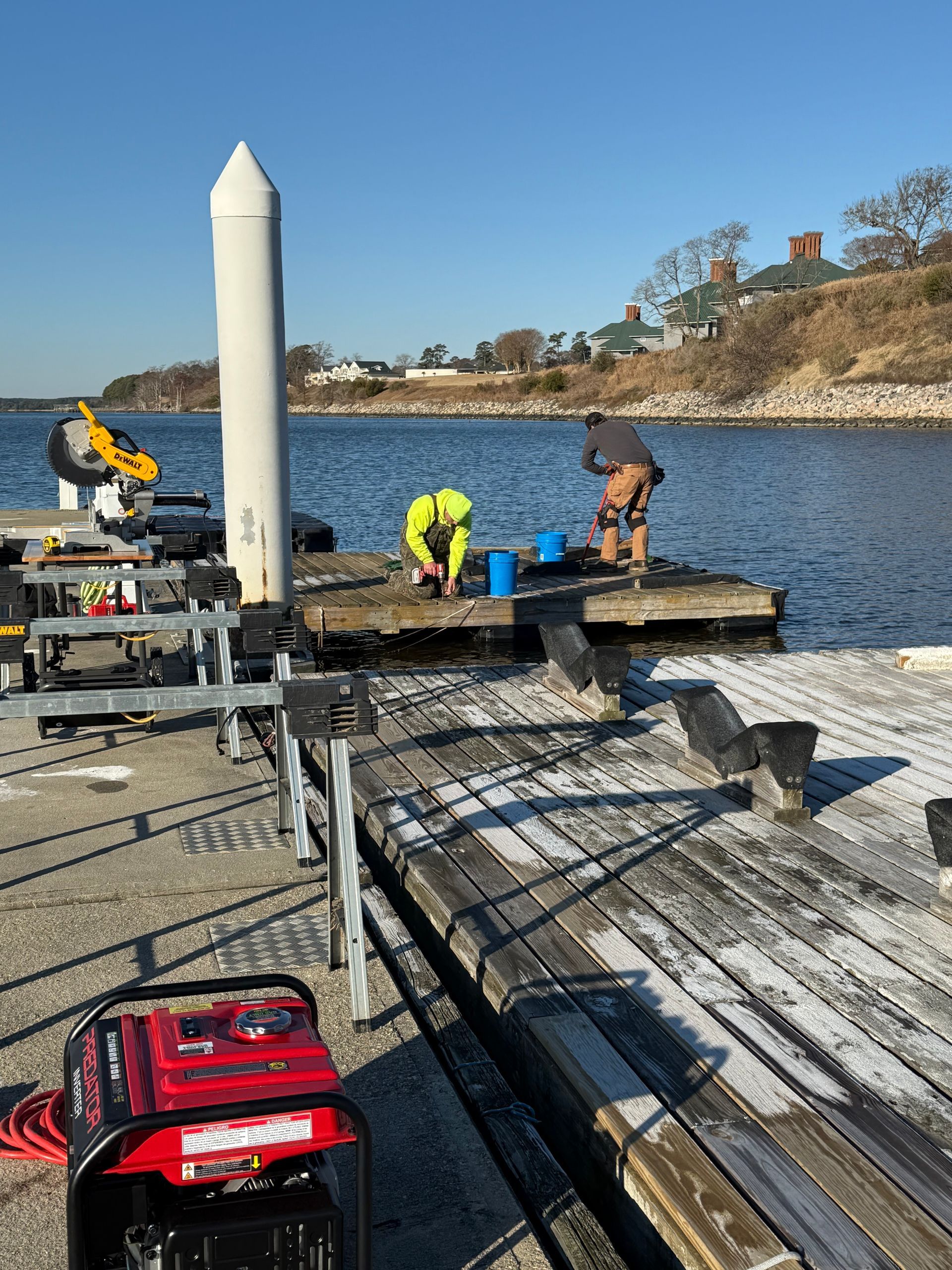 Two workers on a dock repair boards. One wears yellow, the other is hunched over. A generator sits nearby.