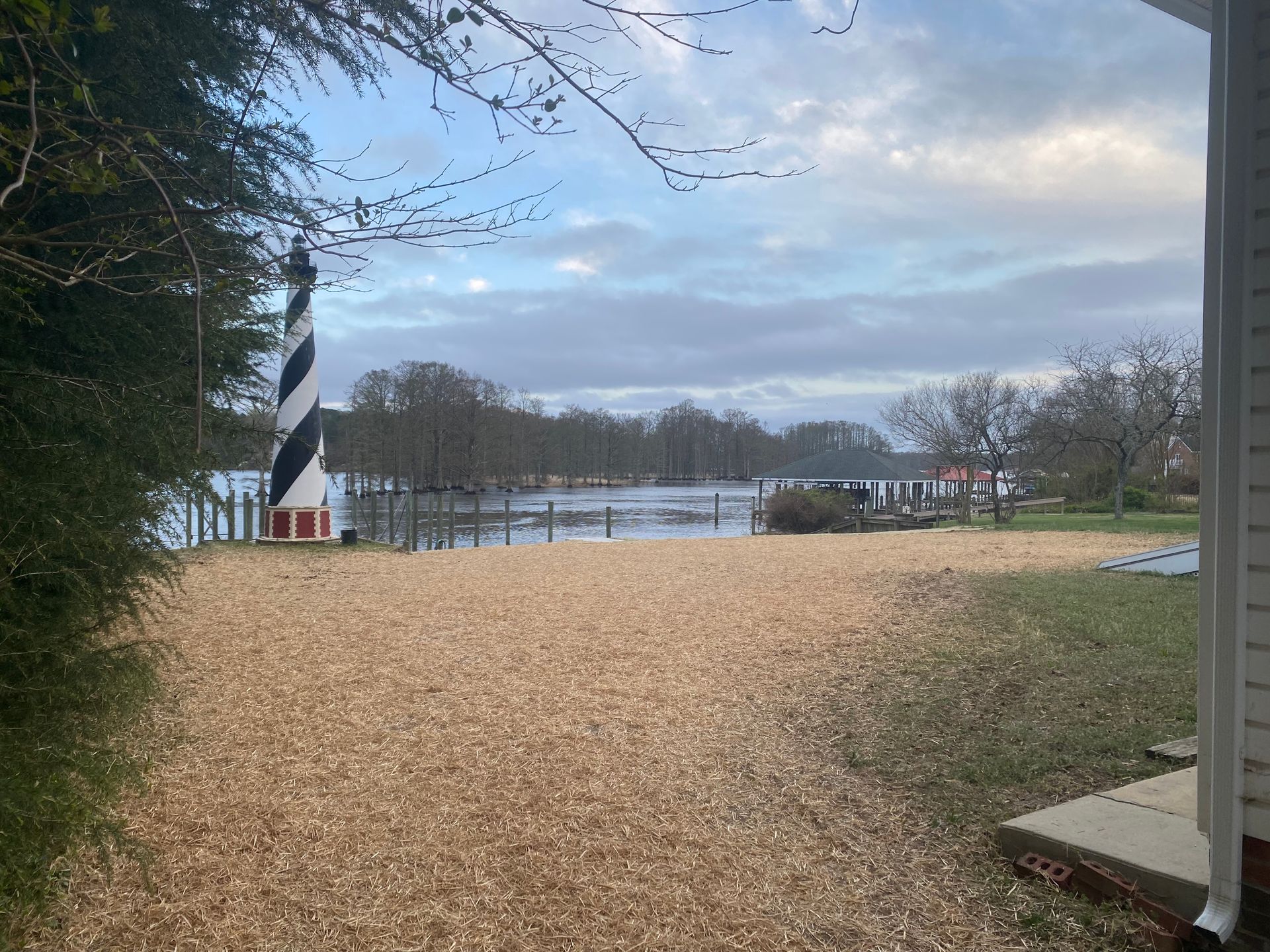 A black-and-white lighthouse stands on a waterfront; a wood-chipped area leads to the water under a cloudy sky.