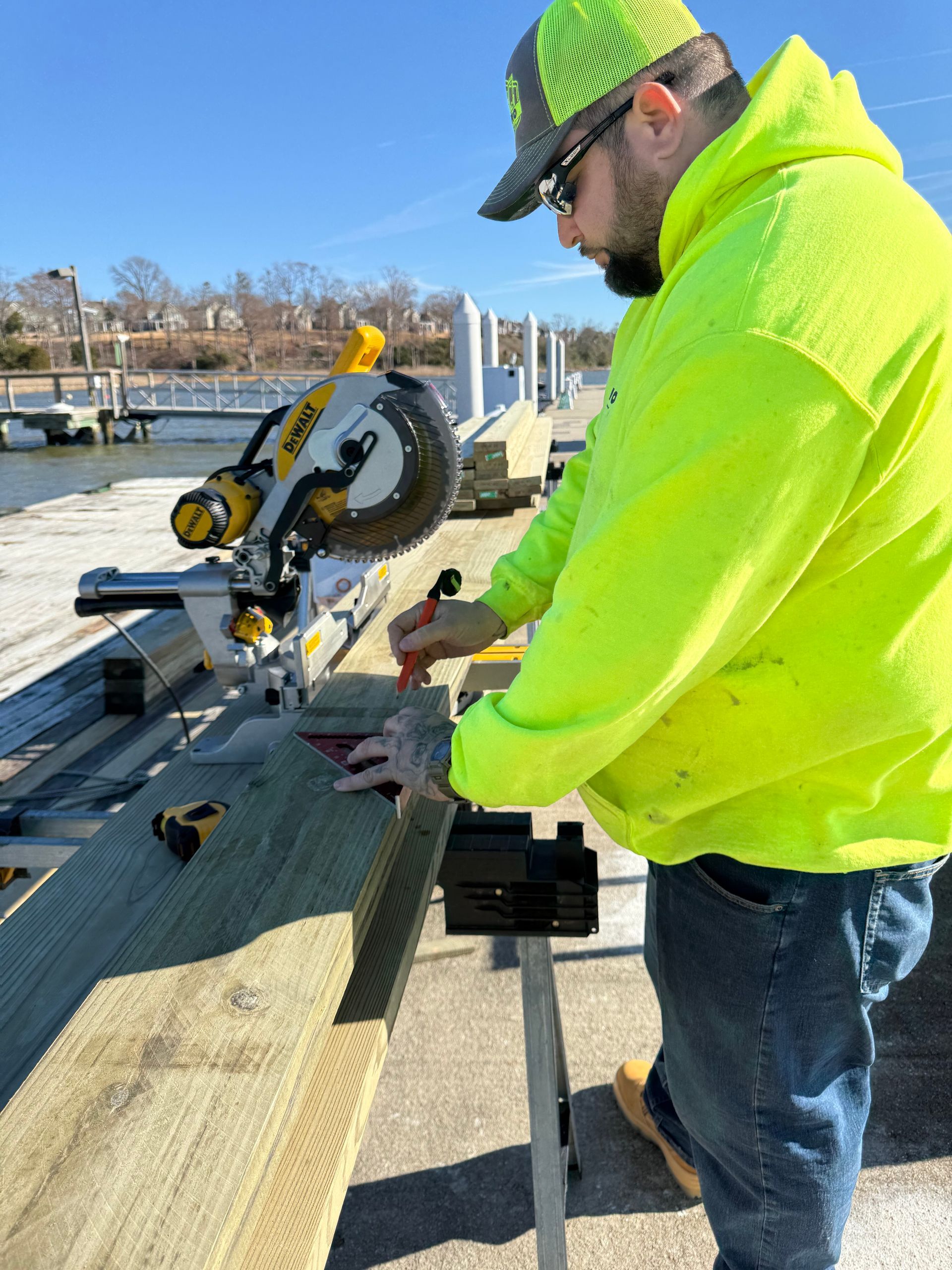 Man in neon yellow hoodie using a circular saw outdoors, cutting wood at a dock.