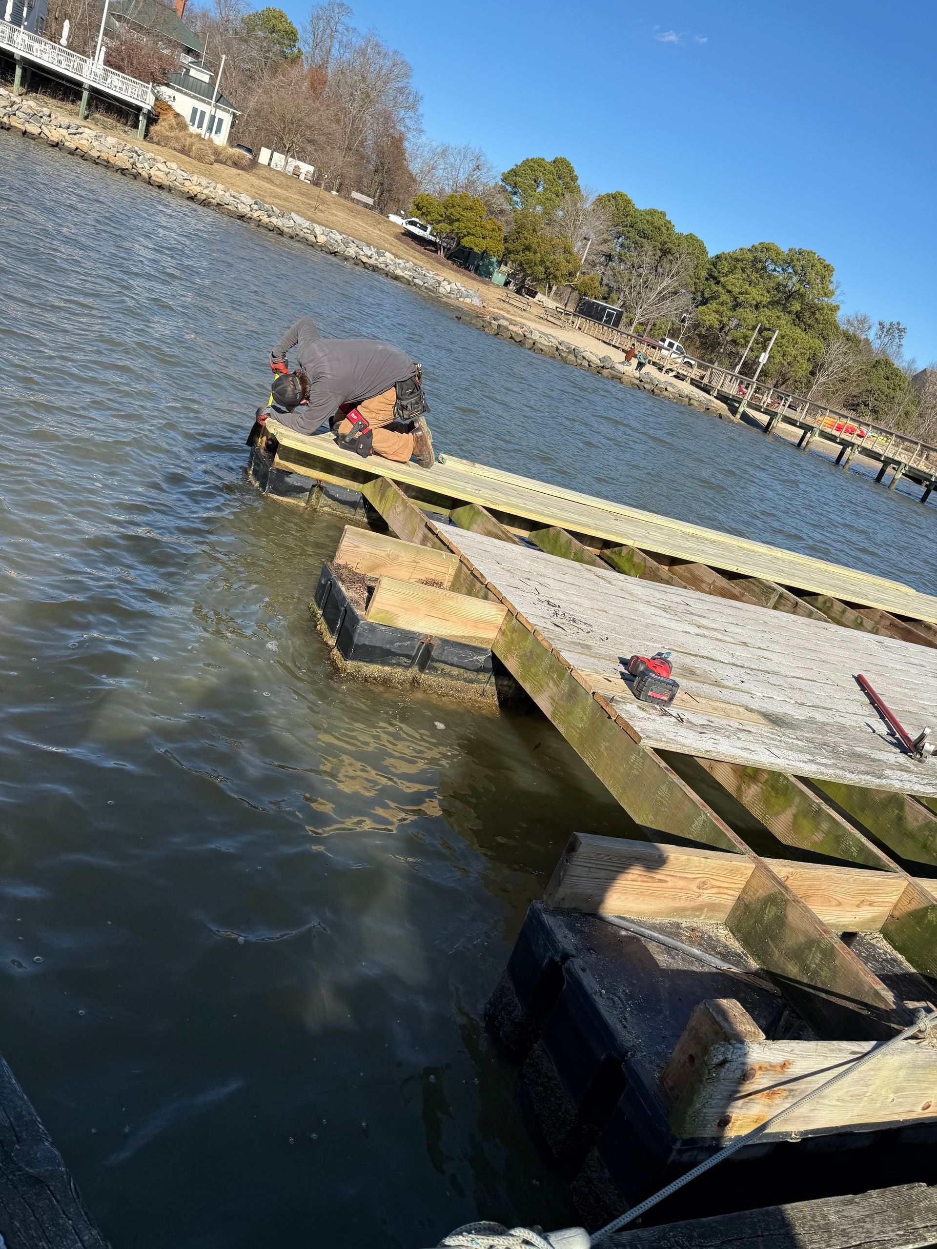 Person working on a weathered dock, near water, with houses on a bank in the background.