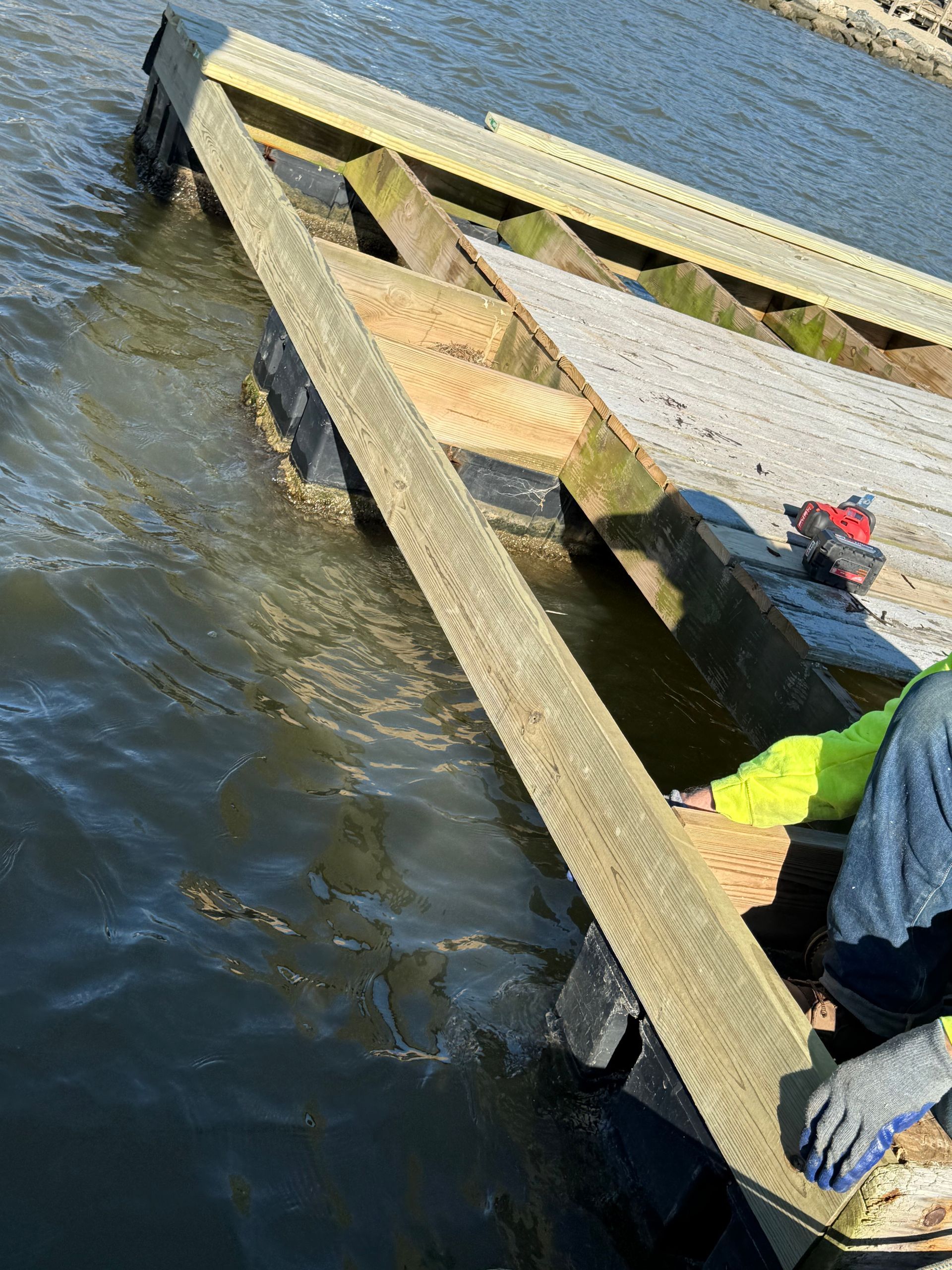 Person building wooden dock over water; yellow and blue work clothes; drill.