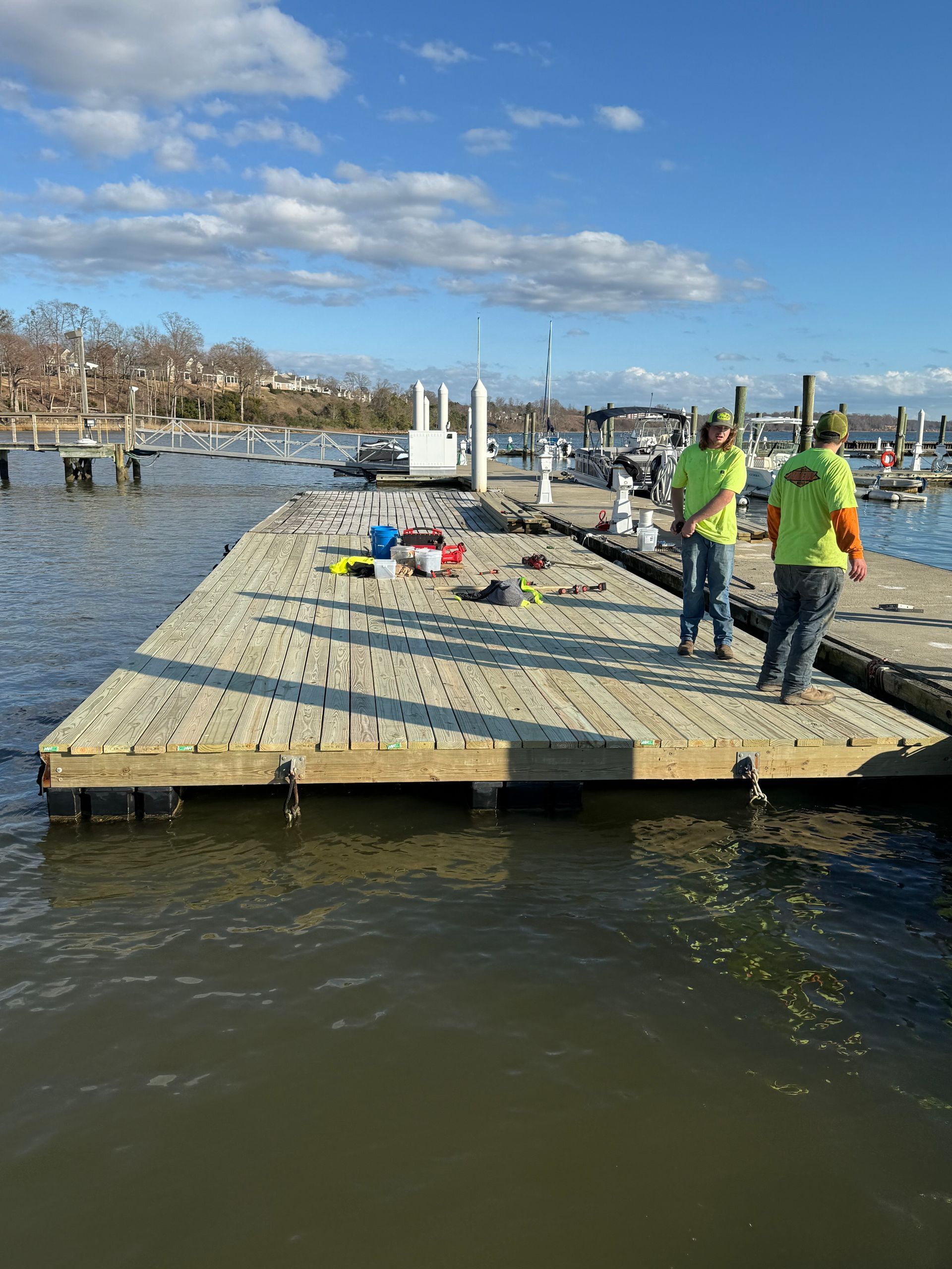 Two workers in neon vests on a dock over water; blue sky, boats and trees in the background.