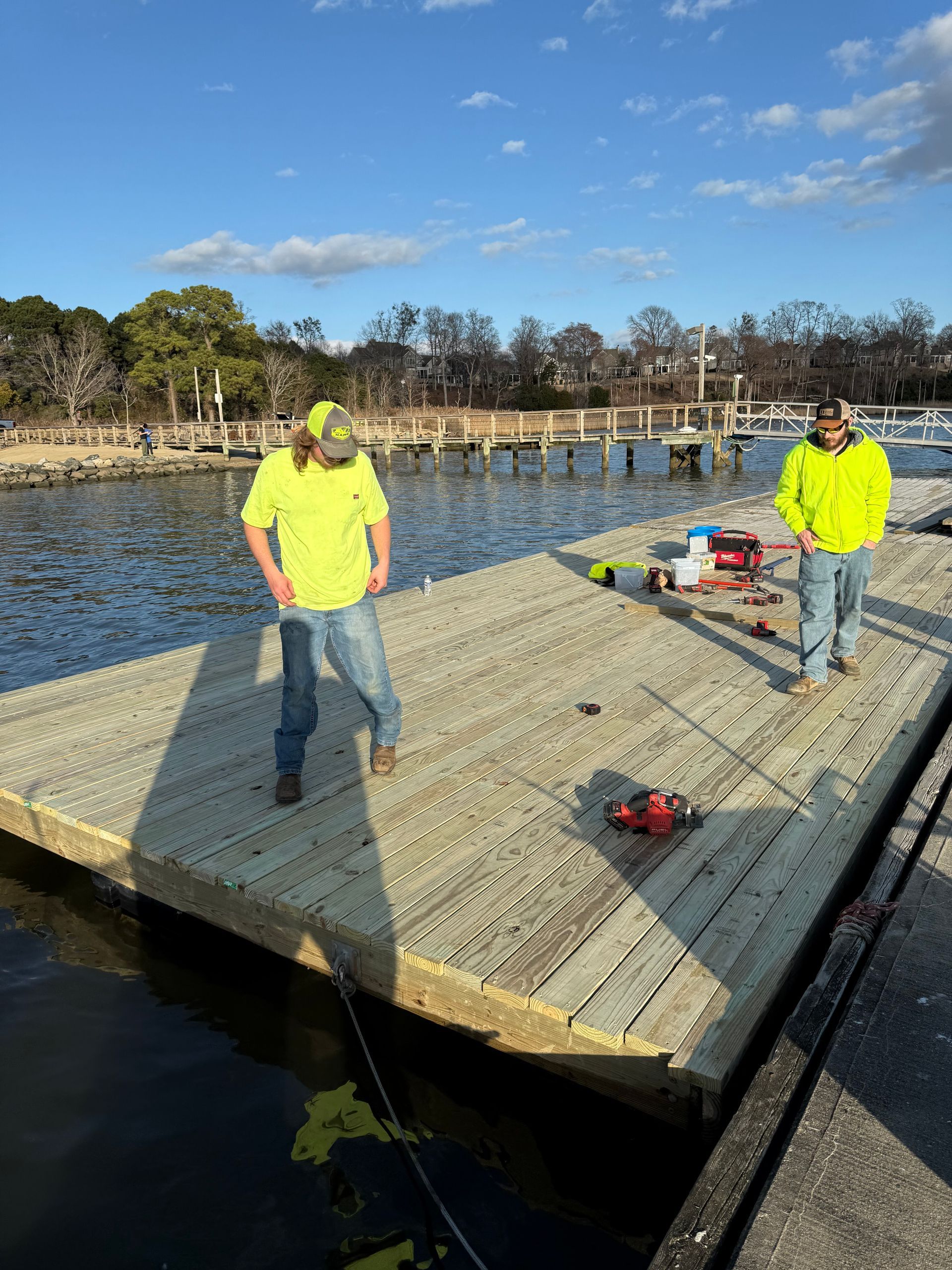Two workers in yellow vests on a floating dock near the water, tools visible; sunny day.