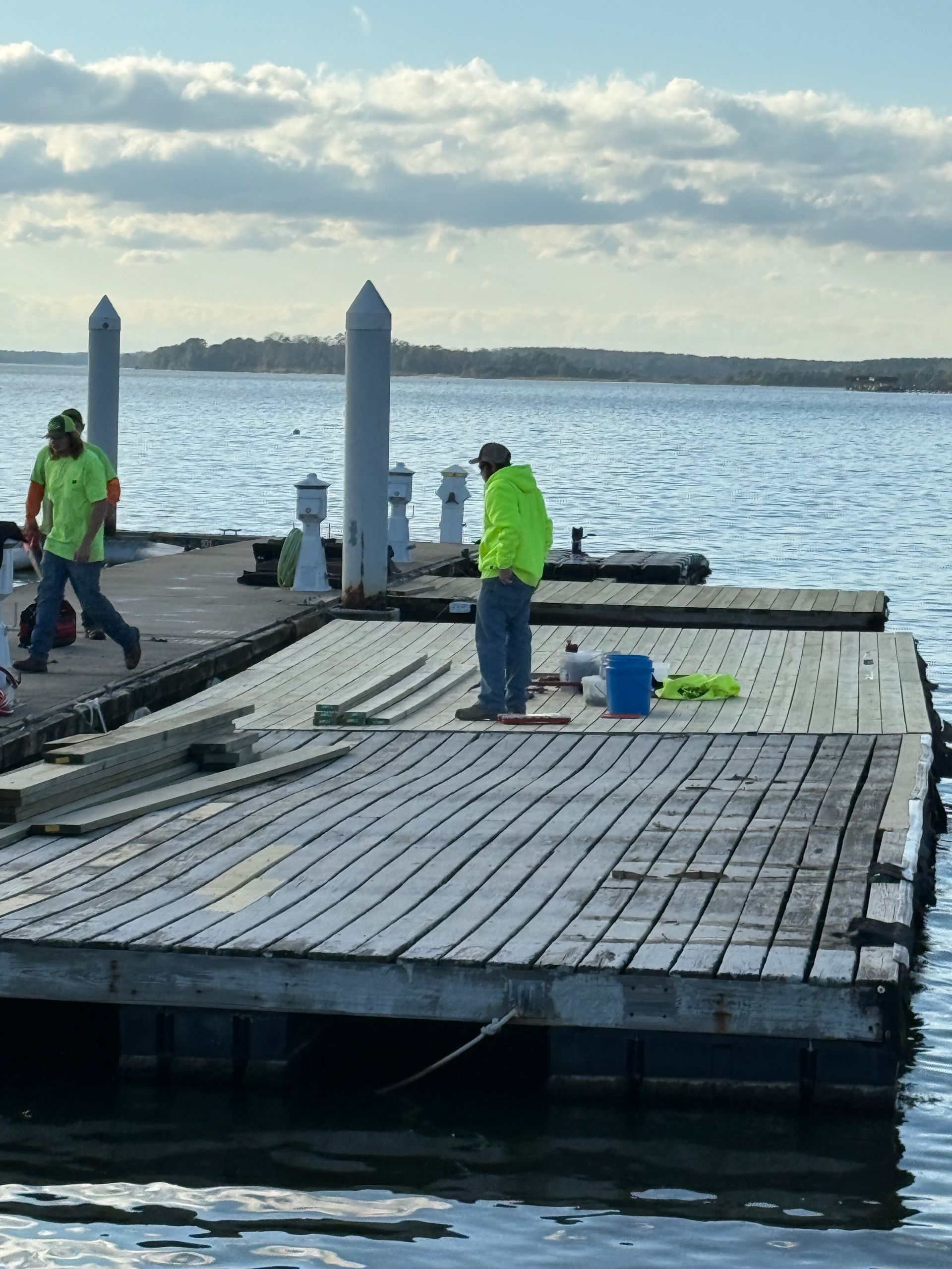 Men in neon green vests working on a wooden dock over water on a cloudy day.