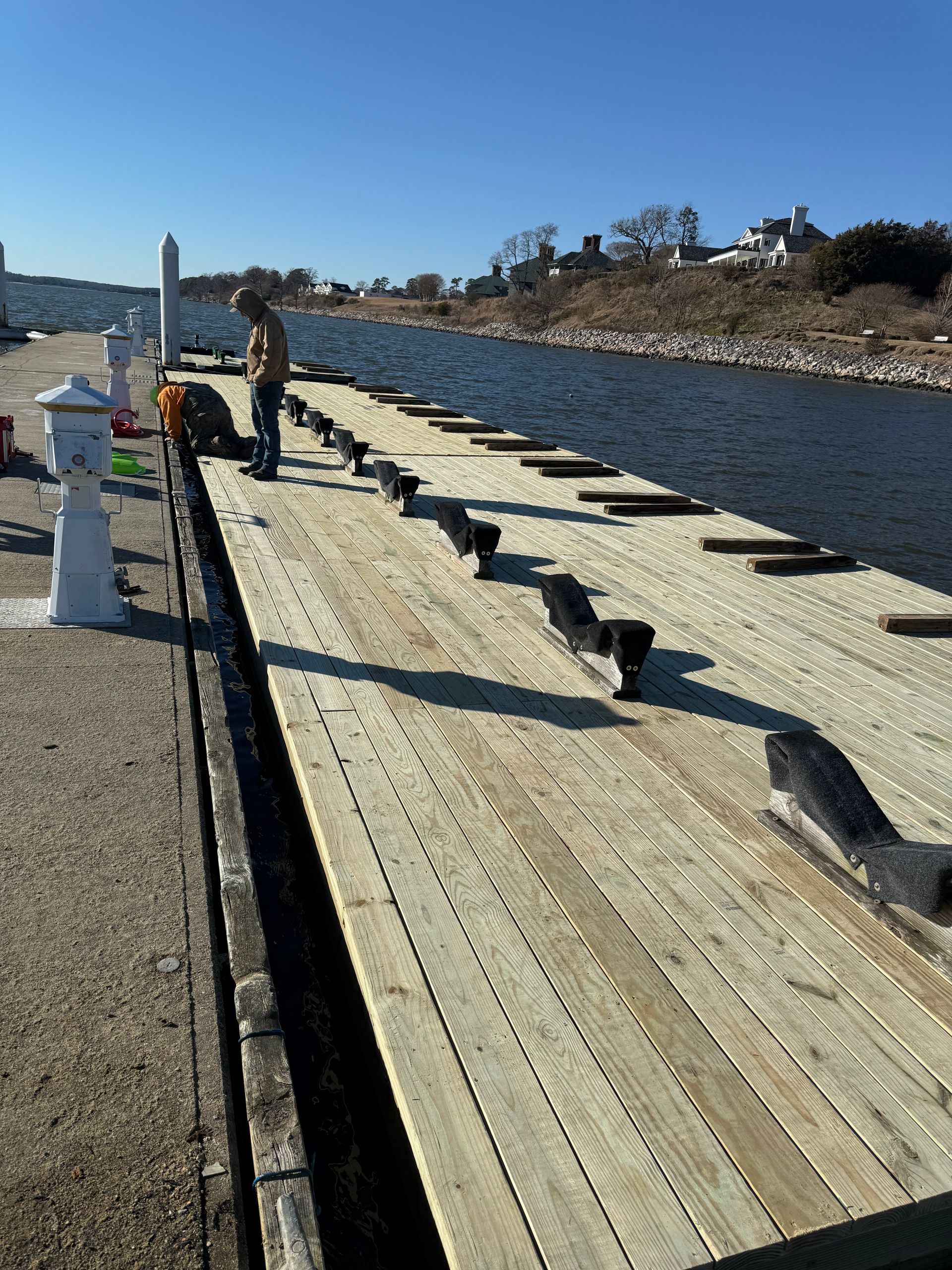 Workers on a wooden dock with new planks, near water. Bright sunlight.