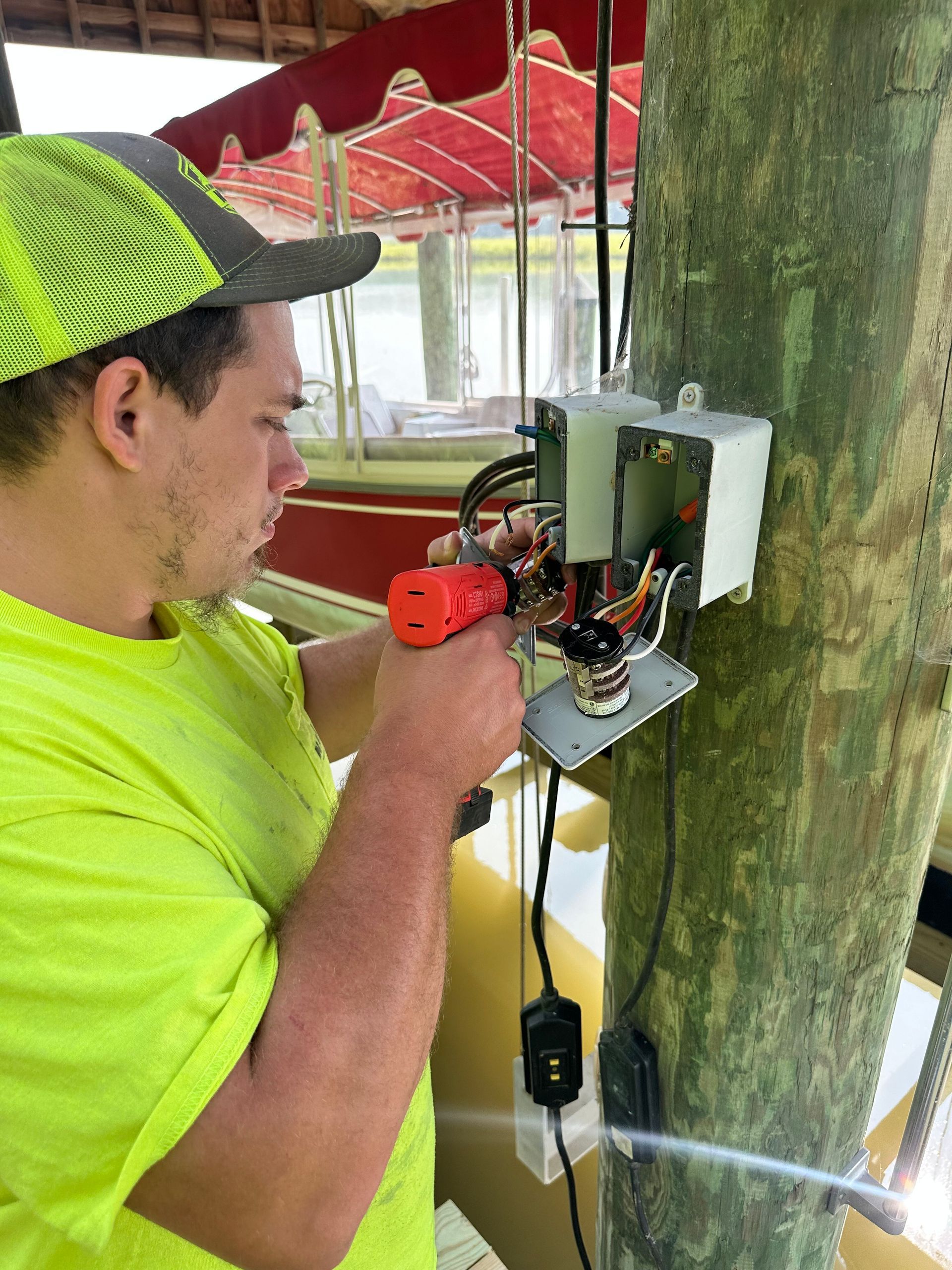 A man in a green shirt and hat uses a power drill on an electrical box on a wooden post.