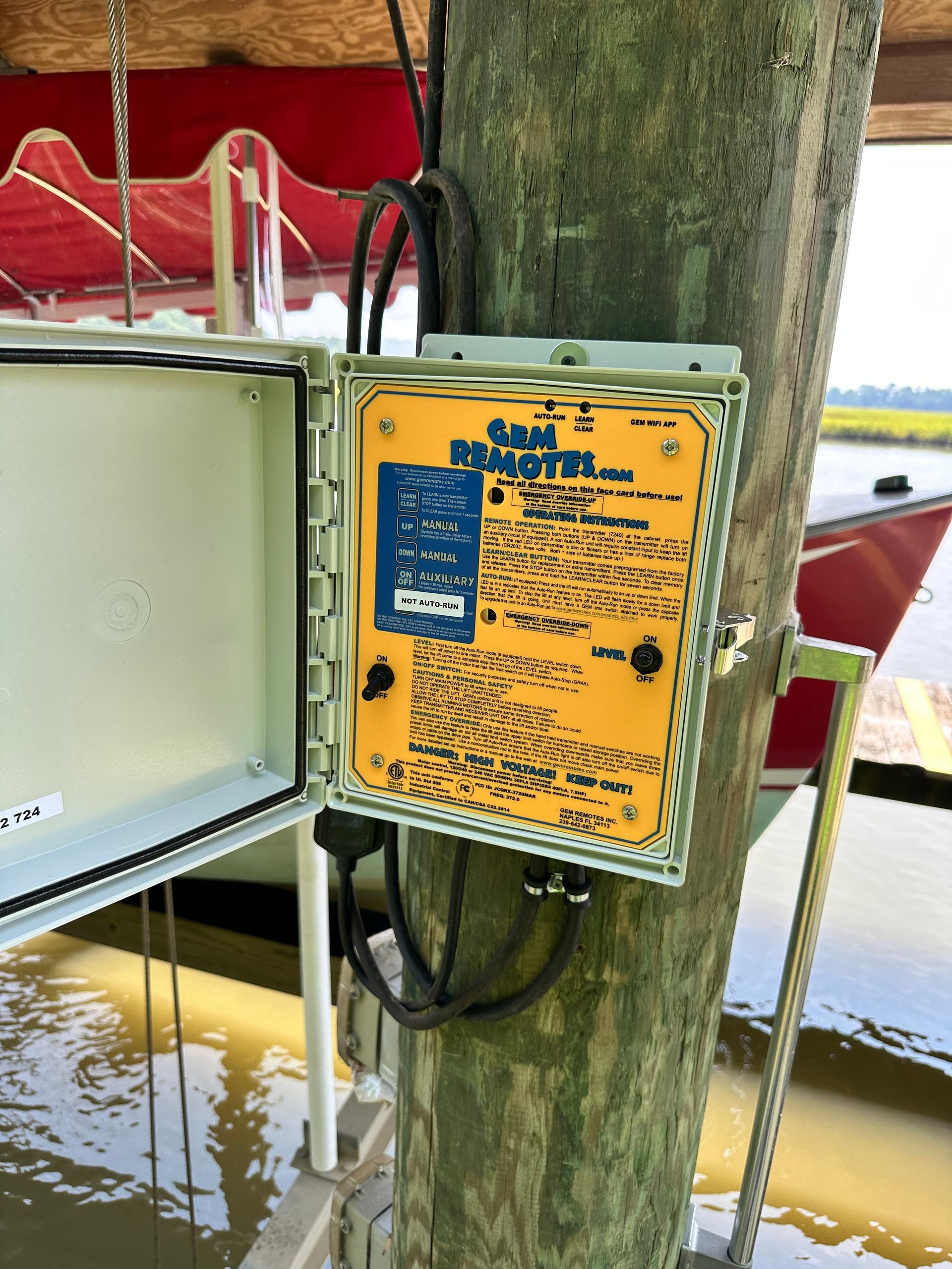 Open electrical box with yellow label attached to a wooden piling over water.