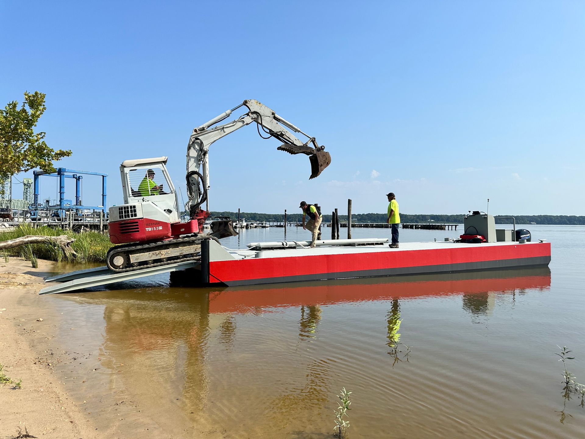 Excavator on a barge, two workers, working at water's edge. Bright day.