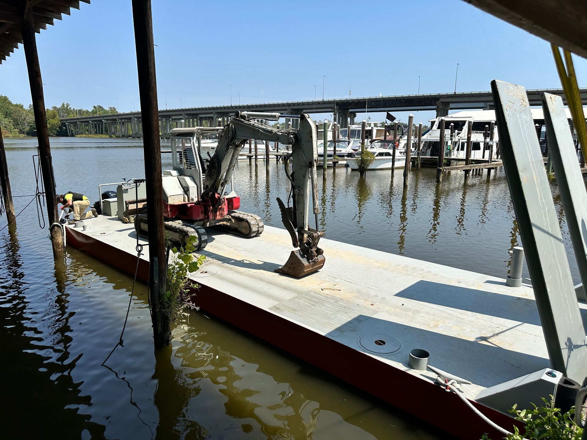 An excavator on a barge dredges a waterway near a marina, under a bridge on a sunny day.