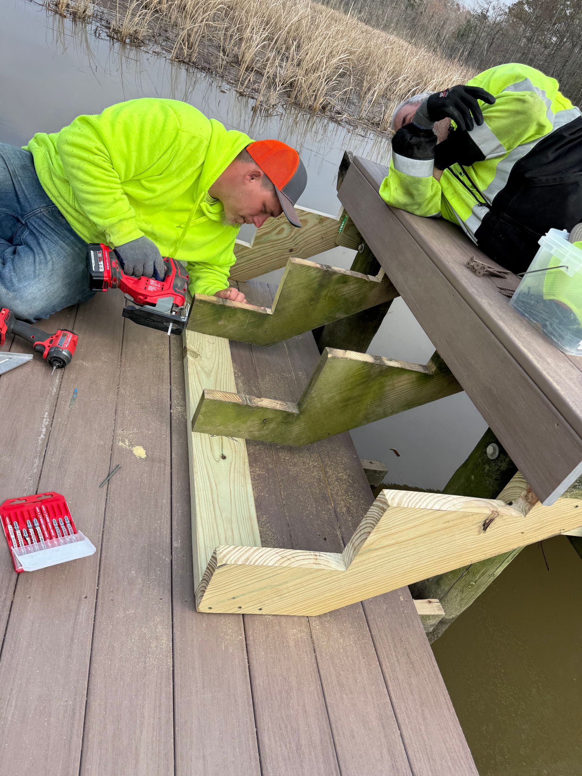 Two people repairing wooden steps on a deck; one uses a saw, both wear safety vests, steps partially submerged.