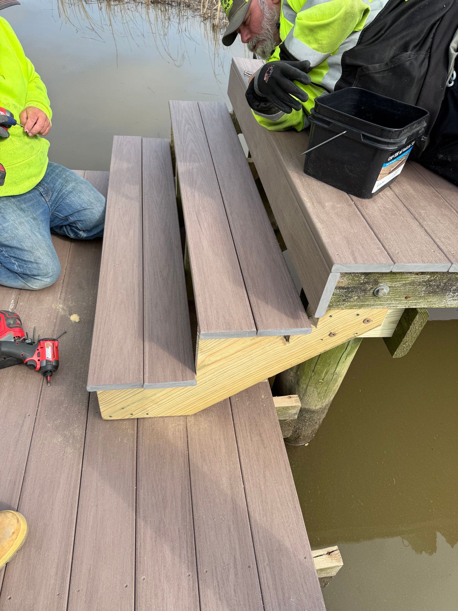 Workers install composite decking on a dock, brown planks, yellow support beams, water visible.