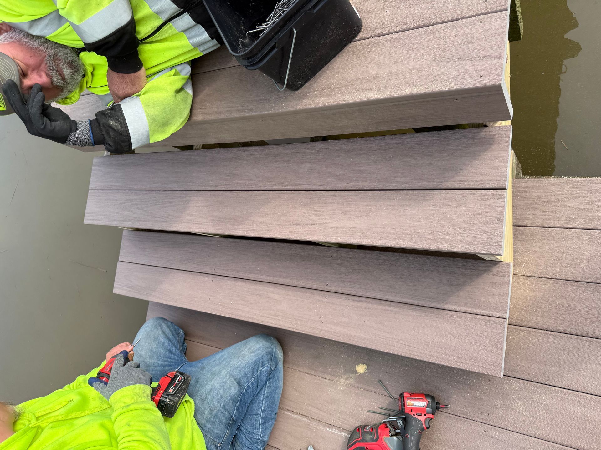Workers installing composite decking steps near water. One drills while the other holds planks.