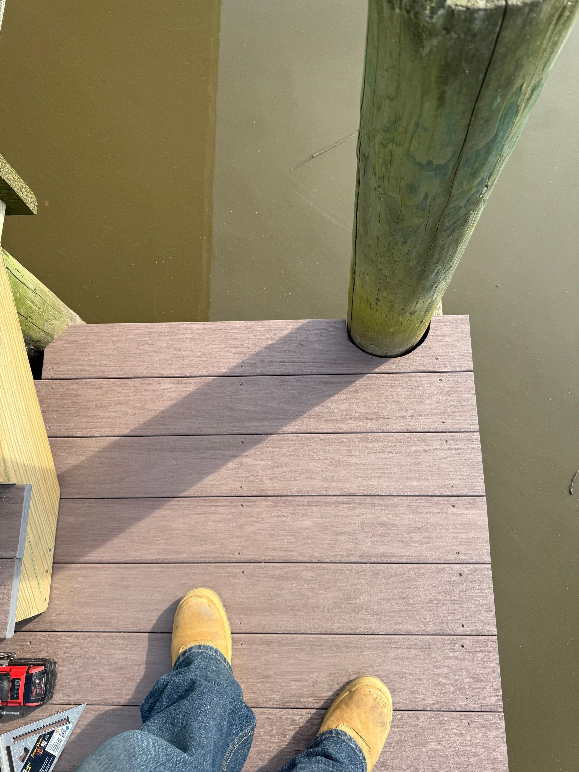 Person standing on a wooden dock by a green-tinted body of water.  Sunlight casts a shadow.