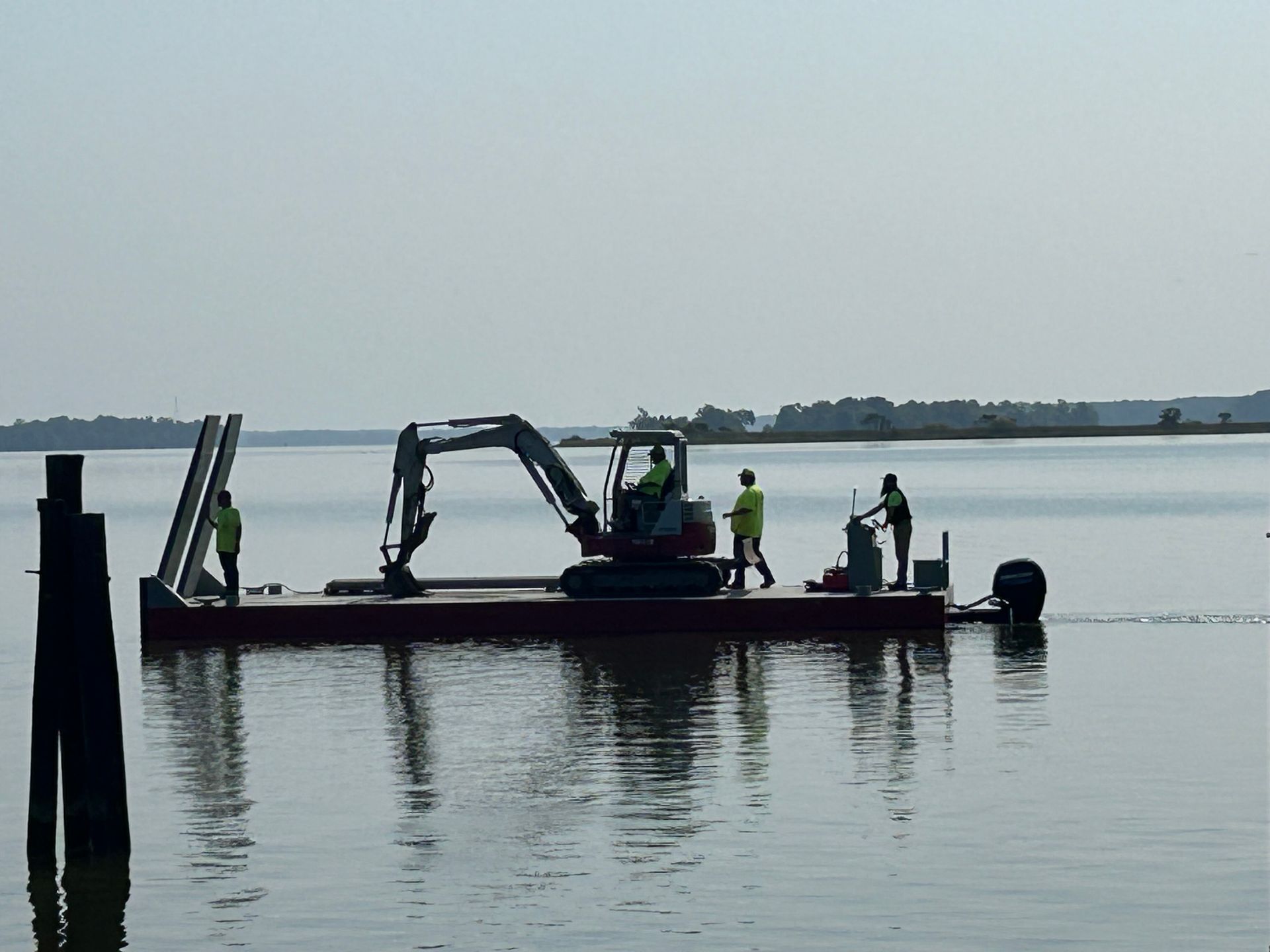 Excavator on a barge dredging in water, with workers in vests, a pier, and land in the background.
