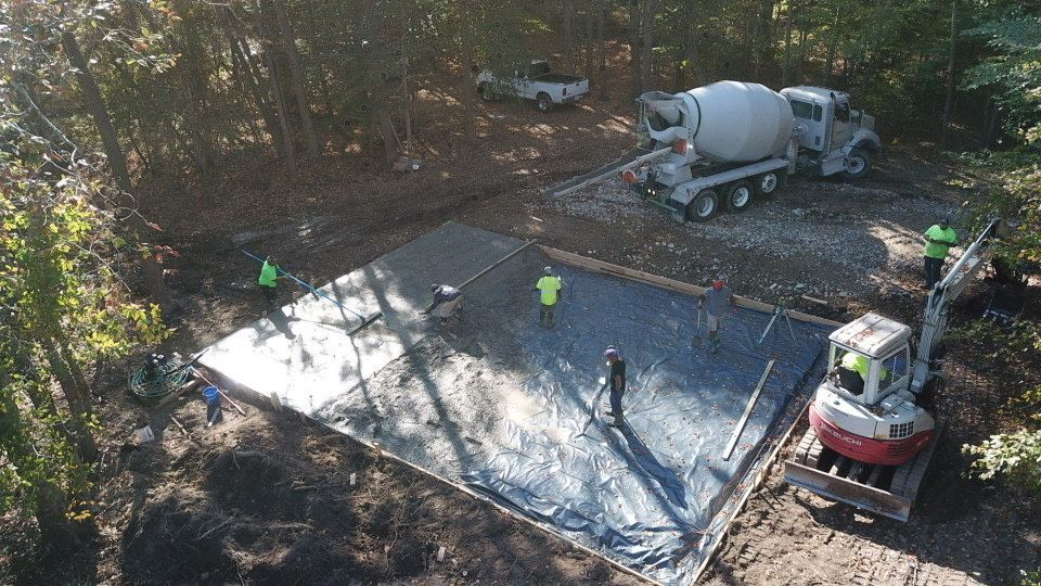 Concrete pour: Cement truck, workers, and mini excavator on a construction site in a wooded area.